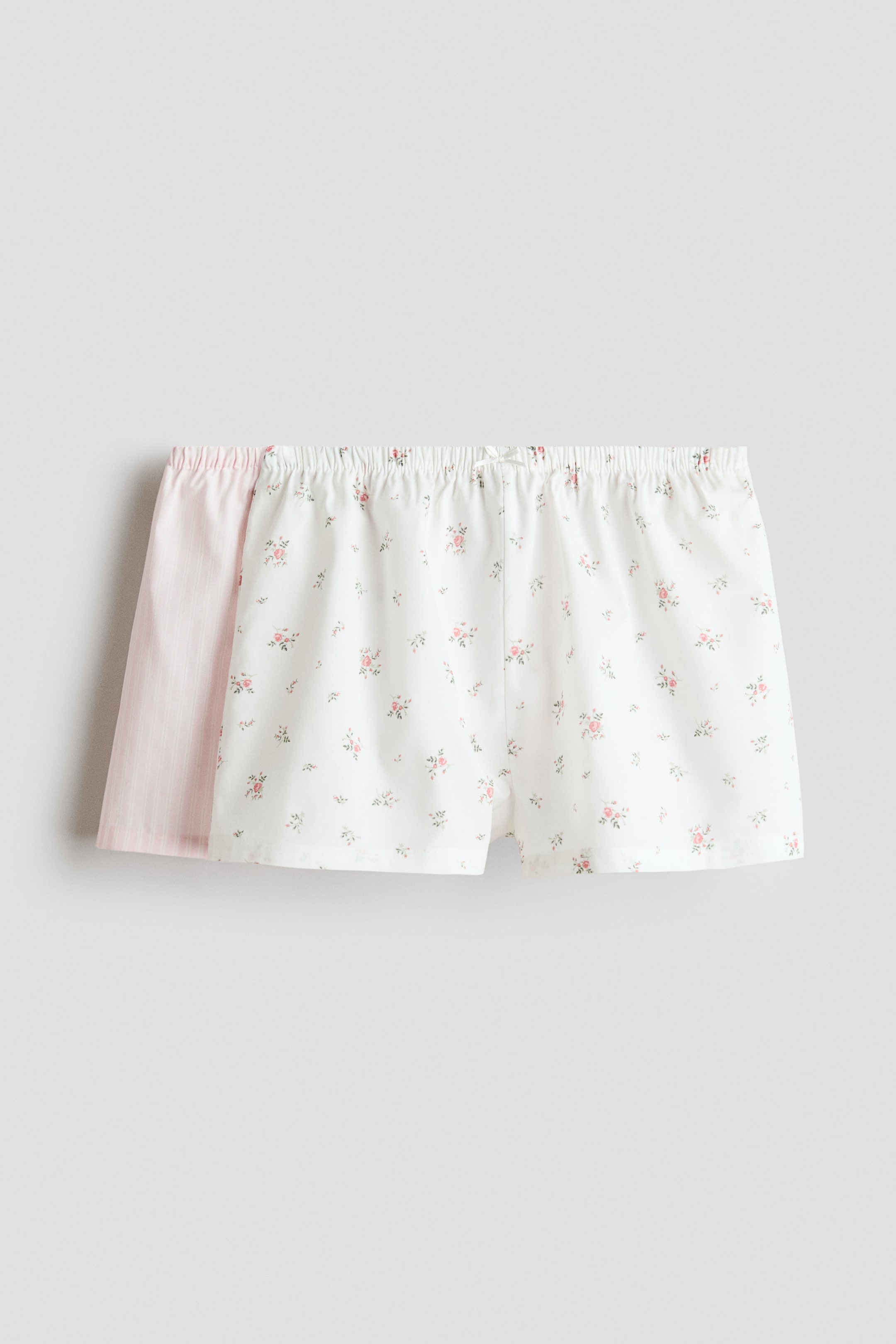 View larger image: Pyjama shorts are showcased, presenting a white pair adorned with a delicate floral pattern facing forward. This pair is crafted with an elasticated, drawstring waist tied into a neat bow and seems to be made from a soft cotton weave, with a pink striped pair partially visible behind it.