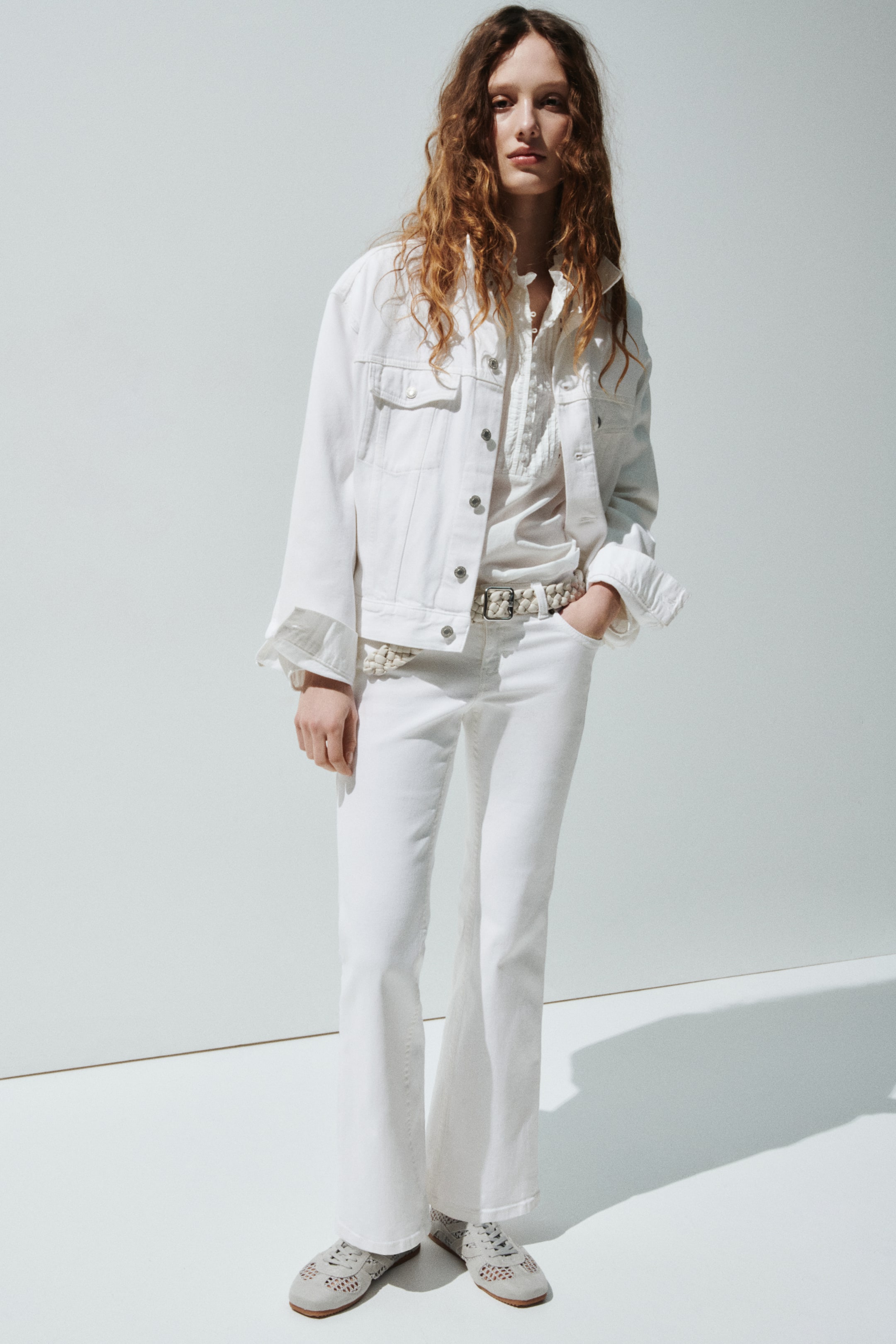Visa större bild: Det vita setet består av en denimjacka med bröstfickor med lock och metallknappar, som bärs öppen över en vit skjorta med en krage med volanger och en plisserad knappslå. Vita utsvängda jeans bärs med ett lätt vävt bälte, och ljusa perforerade platta skor kompletterar den framåtvända looken.