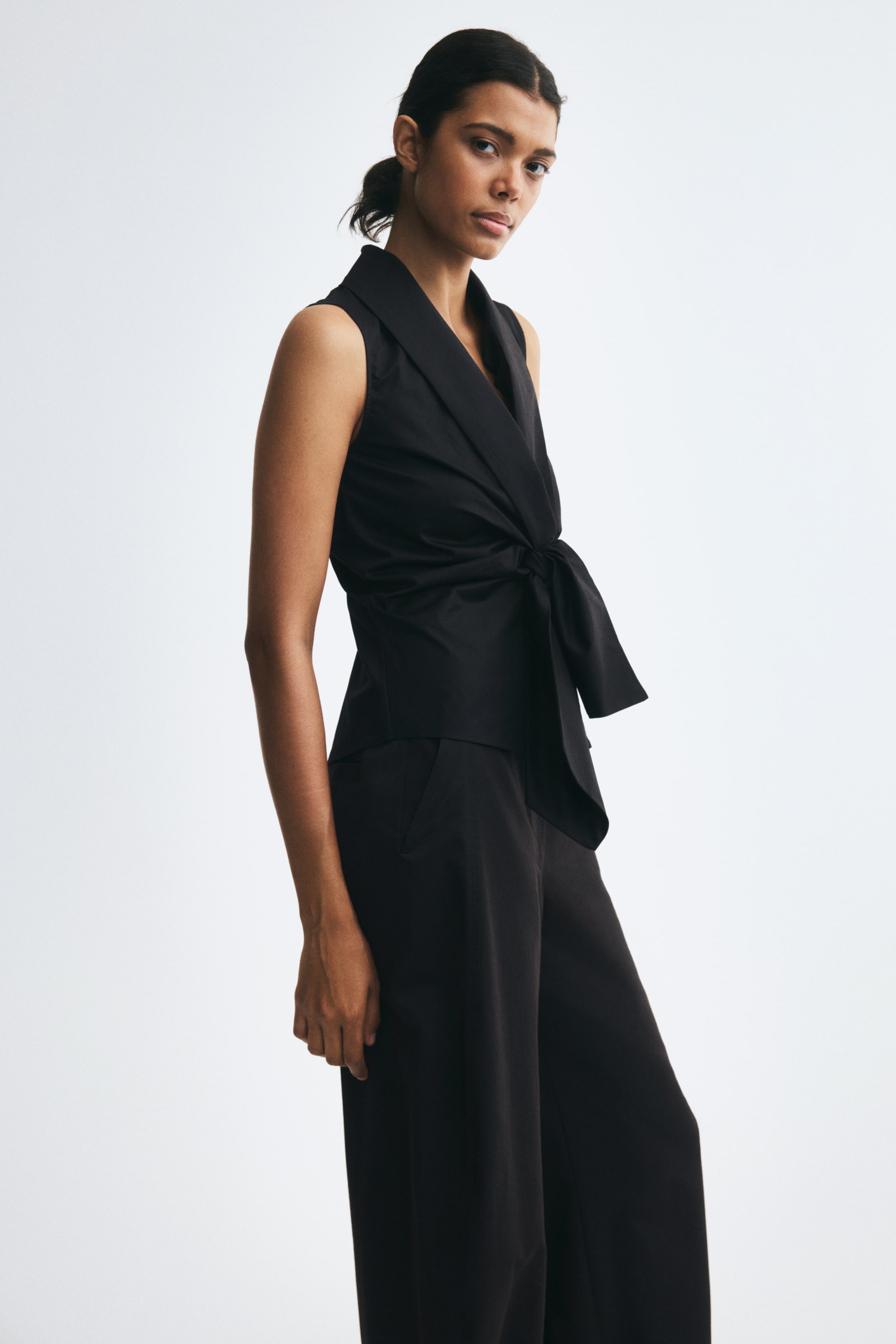 View larger image: A sleeveless black fitted top with a shawl collar and wide ties knotted at the front of the waist is worn by the model. The top is paired with matching black trousers.