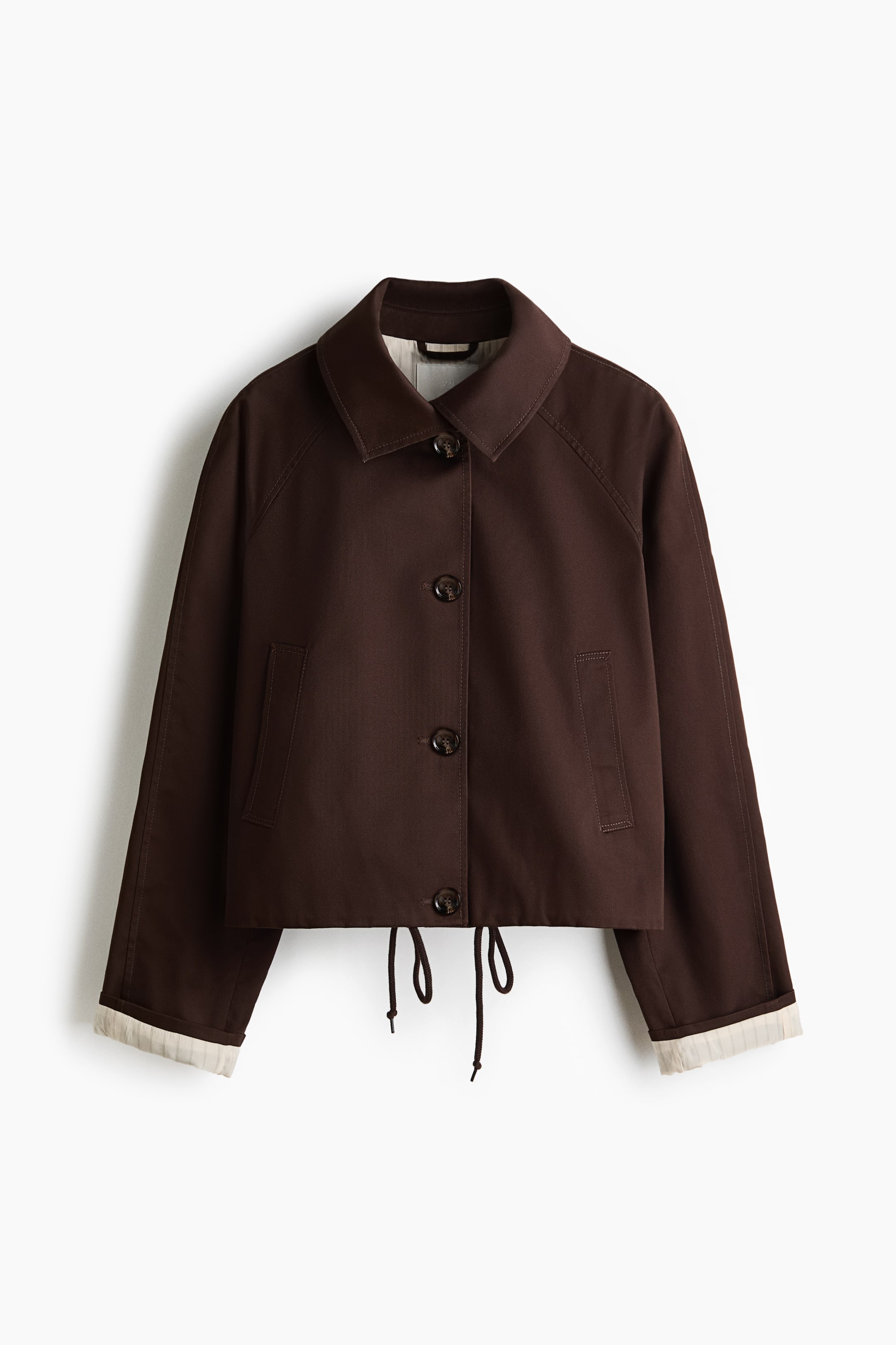 Visualizza immagine più grande: Una giacca corta marrone scuro mostra il davanti, con colletto a punta e chiusura frontale con bottoni. Presenta maniche raglan, due tasche con patta, un orlo con coulisse e polsini ripiegati che rivelano una fodera di colore chiaro.