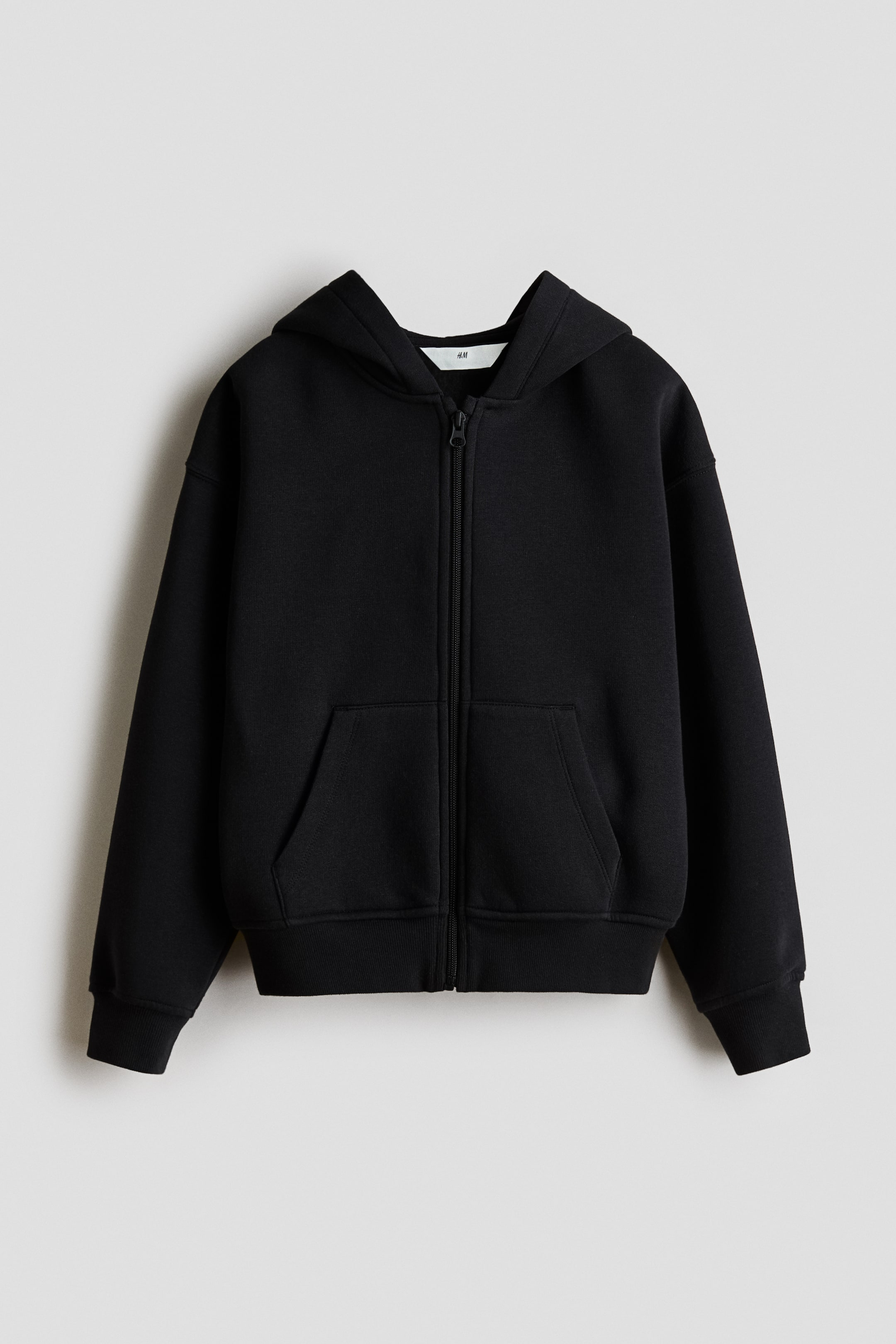 Visa större bild: En svart huvtröja visas framifrån, tillverkad av sweatshirt-tyg. Den har en huva, en central dragkedjestängning, nedhasade axlar, långa ärmar, en framficka i kängurustil och ribbade muddar och fåll.