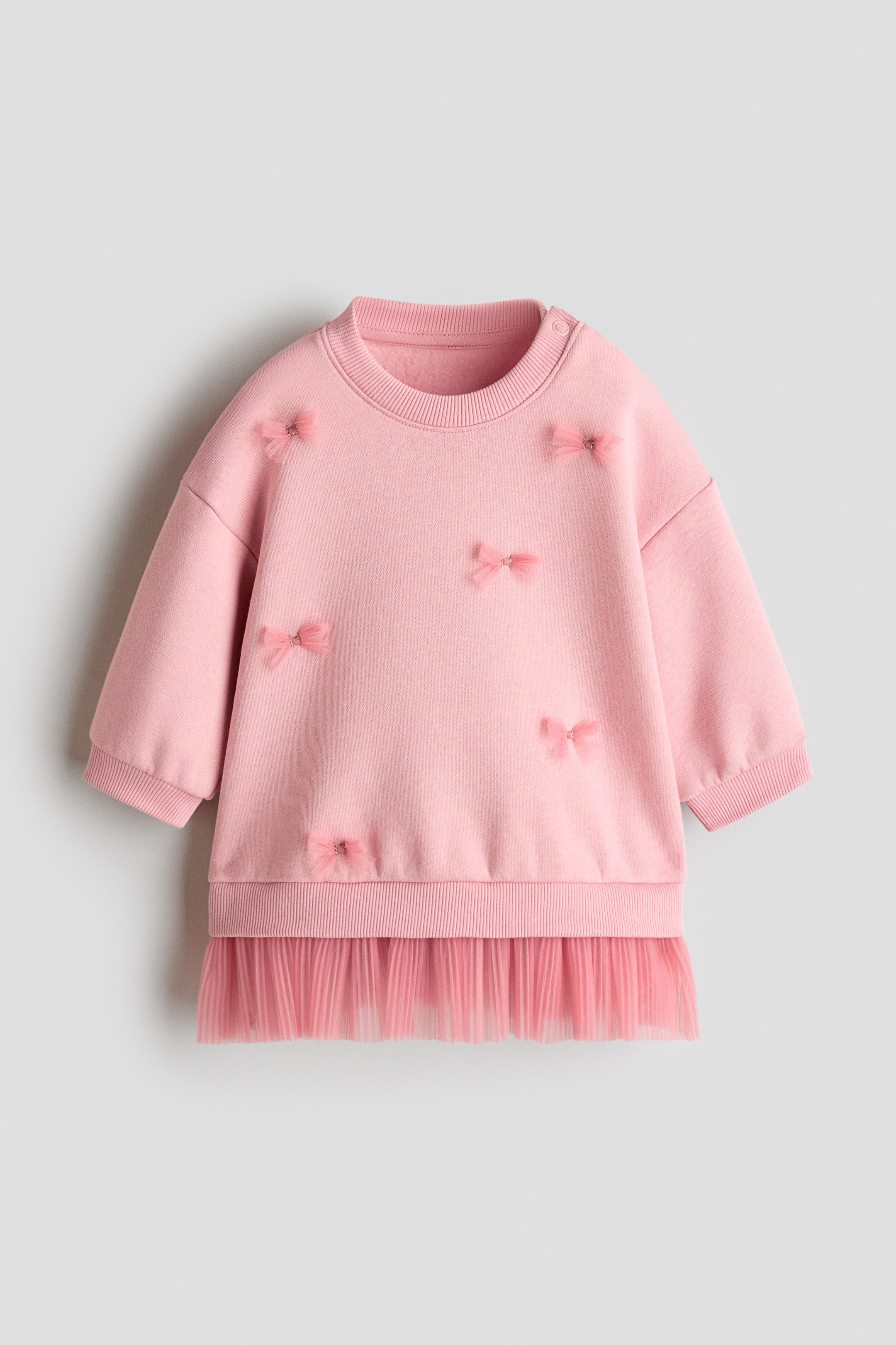 View larger image: A pink sweatshirt dress is shown from the front, featuring a ribbed crew neck with snap buttons on one shoulder and long sleeves with ribbed cuffs. Small sheer pink bows are scattered across the front, and a pleated pink tulle skirt extends from the ribbed hem.