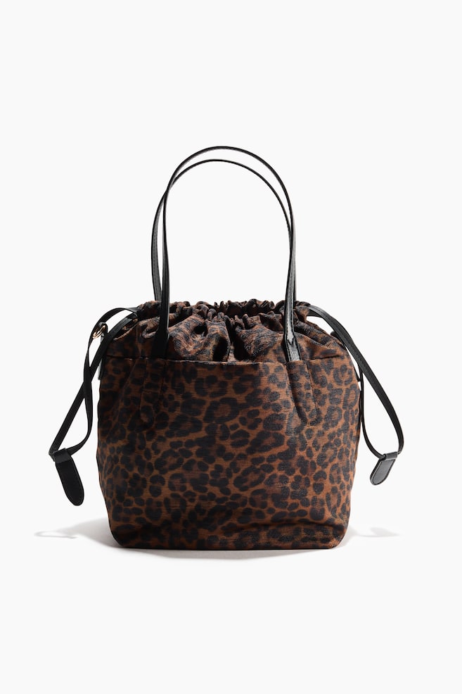 Un bolso de cubo con estampado de leopardo se presenta de frente, ligeramente inclinado hacia la derecha, y cuenta con asas superiores largas y estrechas de color negro con un cierre de cordón fruncido en la parte superior. Una correa de hombro negra, desmontable y ajustable con ganchos de mosquetón, cuelga en su lado izquierdo, y un compartimento abierto es visible en su parte frontal. - 1