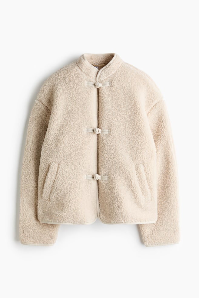 A cream-colored teddy fleece jacket is shown from a front-facing view, featuring a stand collar and three fabric knot button closures down the front. The jacket is designed with a relaxed fit, dropped shoulders, long sleeves, and two slanted welt pockets. - 1