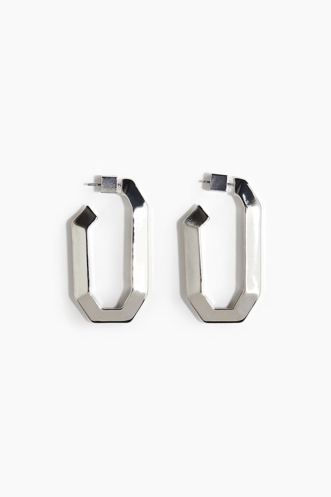 A pair of silver-toned, elongated, multi-faceted hoop earrings is presented front-facing. Each earring is featuring a geometric design with sharp angles and a visible post-back closure. - 1