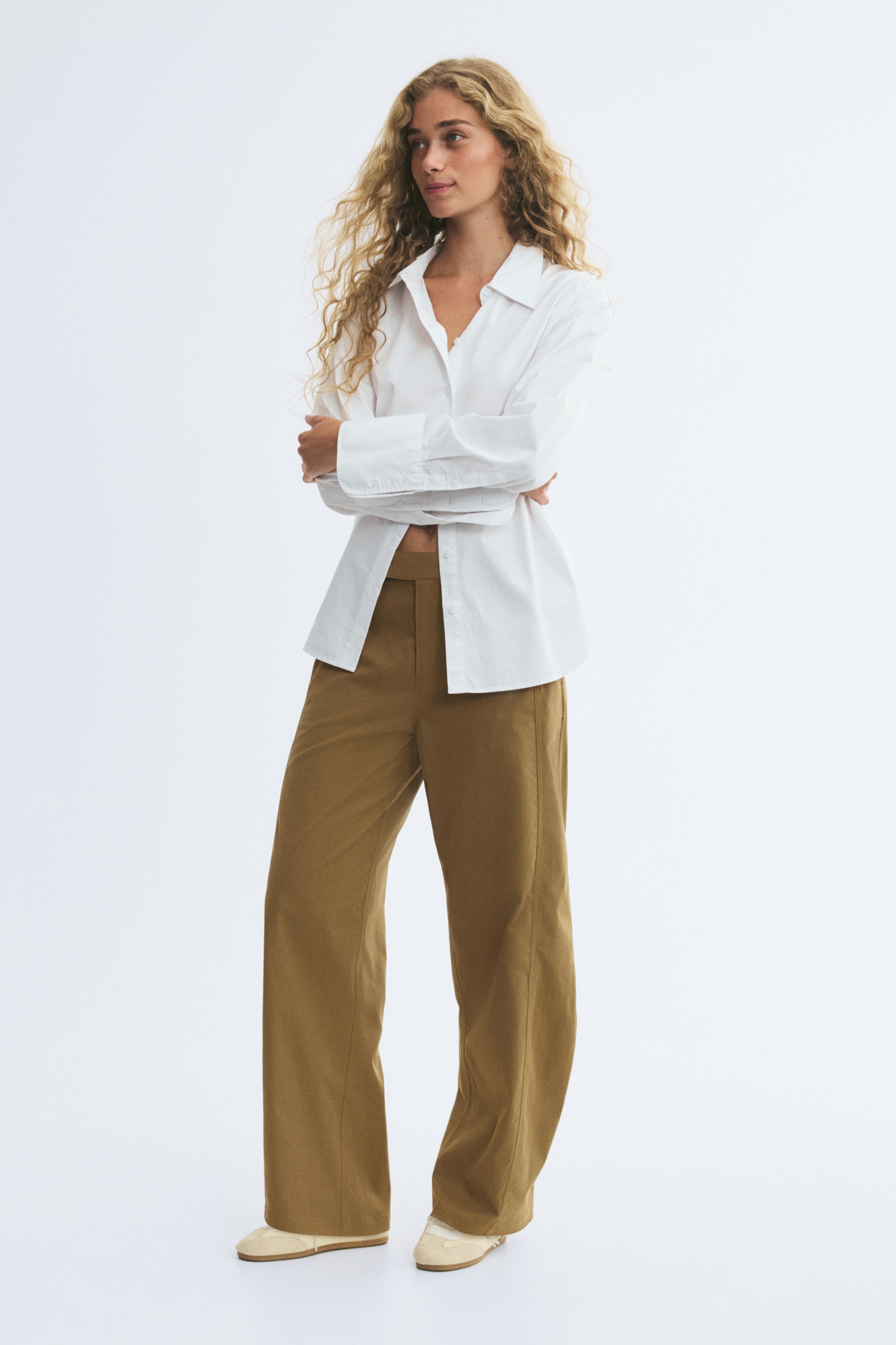 View larger image: Twill pants in a golden-brown shade, featuring a barrel-leg silhouette with forward-facing side seams and an extended waistband, are worn by the model. The pants are styled with a white long-sleeved button-down shirt and beige shoes.