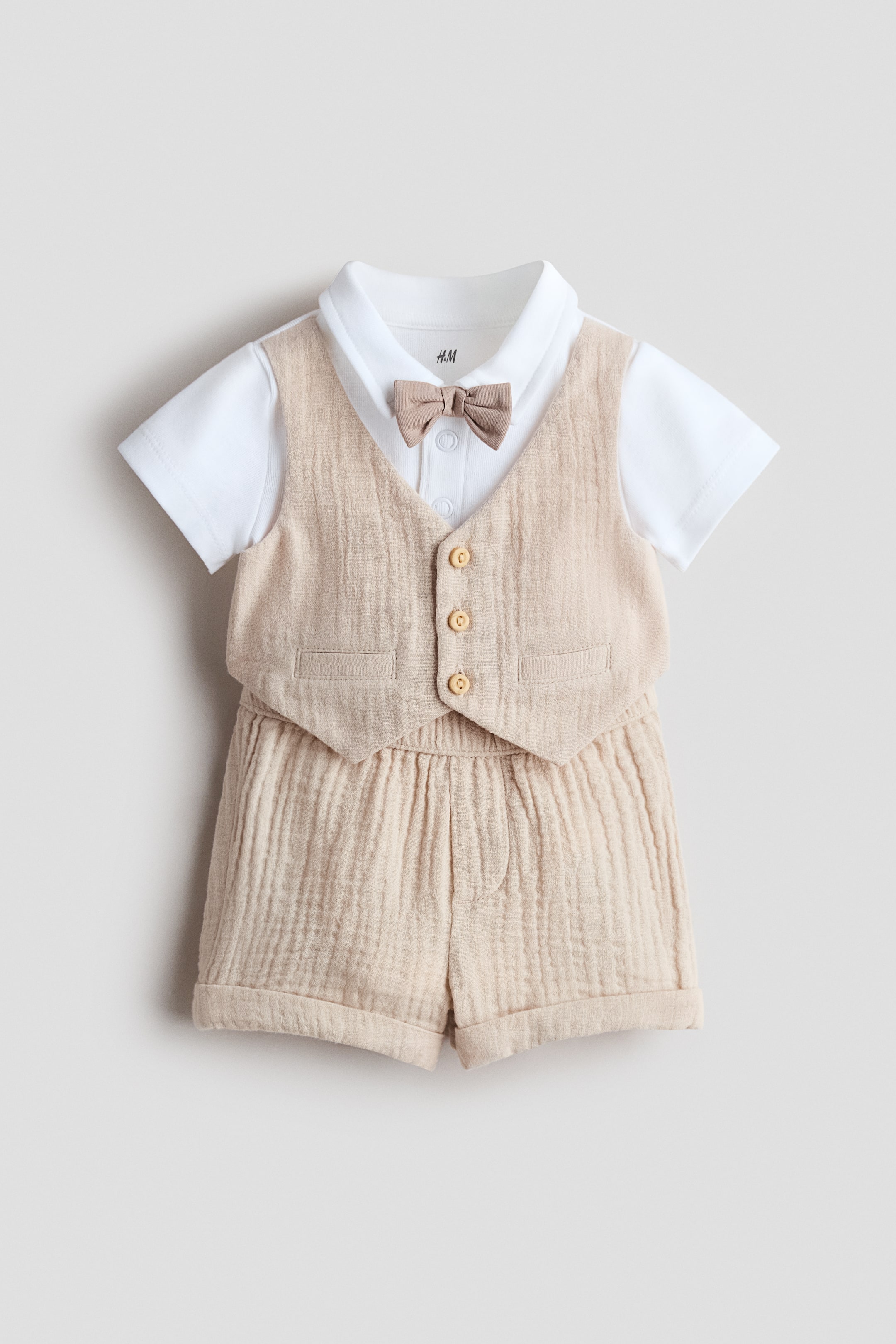 Ingrandisci l'immagine: Un completo a due pezzi per neonati presenta una maglietta bianca a maniche corte sotto un gilet in mussola beige stropicciata e pantaloncini coordinati. Il gilet è progettato con tre bottoni in legno sul davanti e due piccole tasche con patta, con un papillon beige che adorna il colletto della maglietta. I pantaloncini sono progettati con un elastico in vita e orli risvoltati.