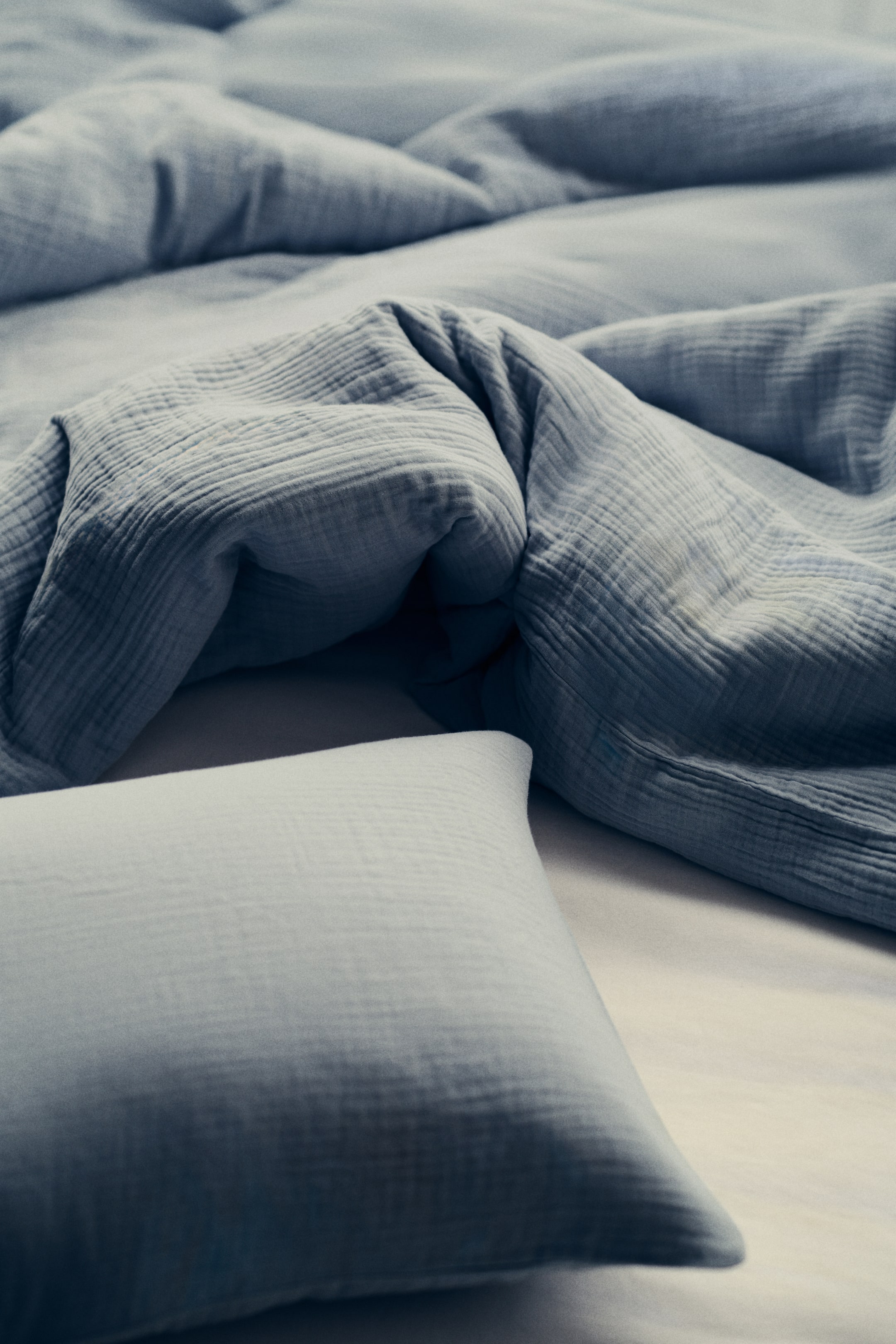 이미지 확대 보기: 파란 회색의 주름진 거즈 이불이 부드러운 질감을 드러내며 드리워져 있습니다. 앞쪽에는 약간 오른쪽으로 기울어진 동일한 색상의 직사각형 베개가 놓여 있습니다.