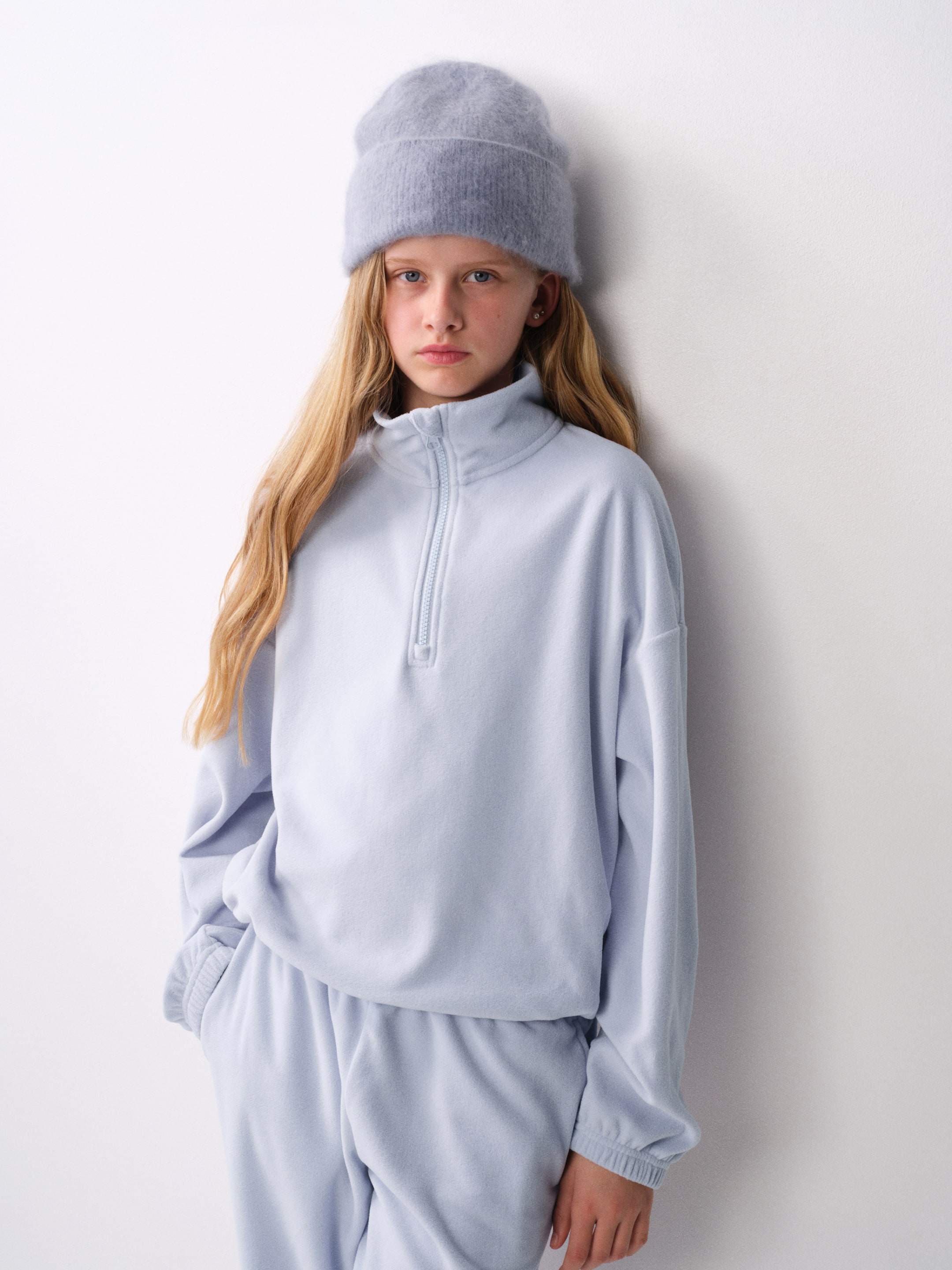 Grösseres Bild ansehen: Ein hellblaues Activewear-Top aus Fleece wird von vorne gezeigt. Es verfügt über einen Stehkragen mit halbem Reissverschluss, überschnittene Schultern und lange Ärmel mit elastischen Bündchen. Das Top wird in einer lockeren, regulären Passform getragen und mit einer passenden Fleecehose und einer Strickmütze kombiniert.