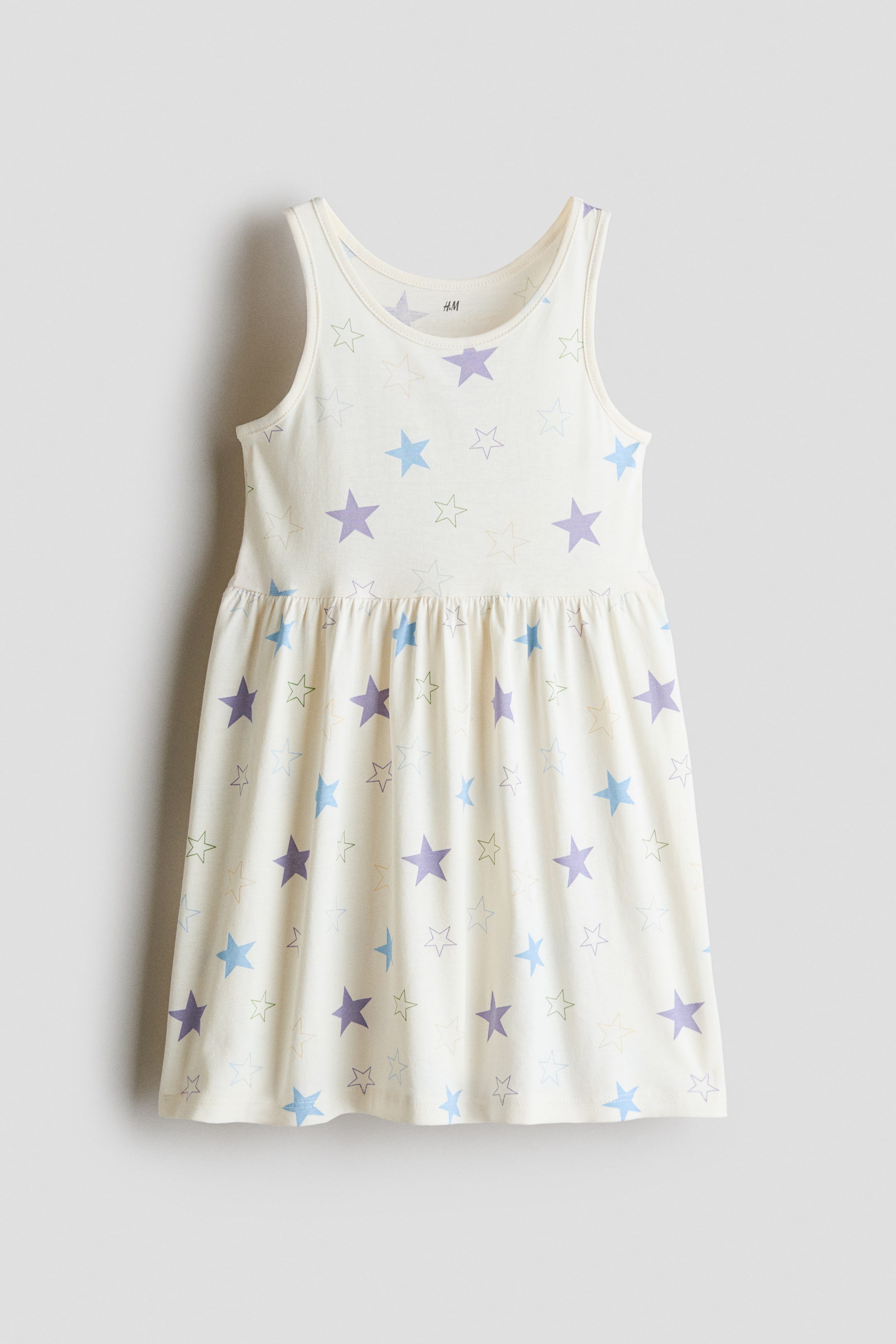 View larger image: A sleeveless, cream-coloured dress is shown from the front, featuring an all-over pattern of solid and outlined stars in pastel shades of purple, blue, and light green. The dress features a scoop neckline, wide shoulder straps, and a gathered waistline that flows into a flared skirt.