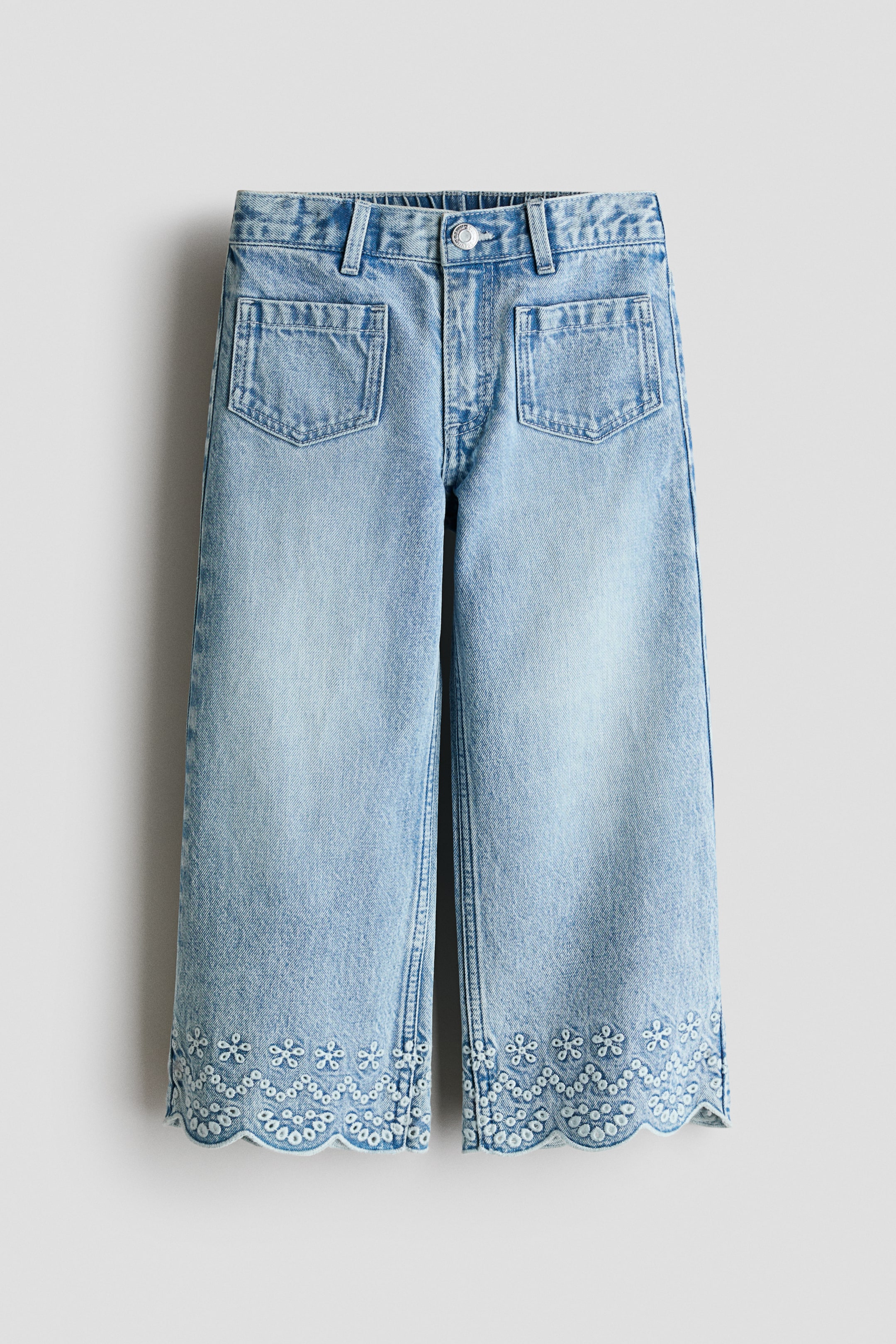 Agrandir l'image: Jean en denim délavé clair face avant, présentant une taille haute avec passants de ceinture, une fermeture à bouton unique, une braguette zippée et deux grandes poches plaquées rectangulaires à l'avant. Les jambes larges et raccourcies se terminent par un ourlet festonné orné de broderies anglaises blanches à motif floral.