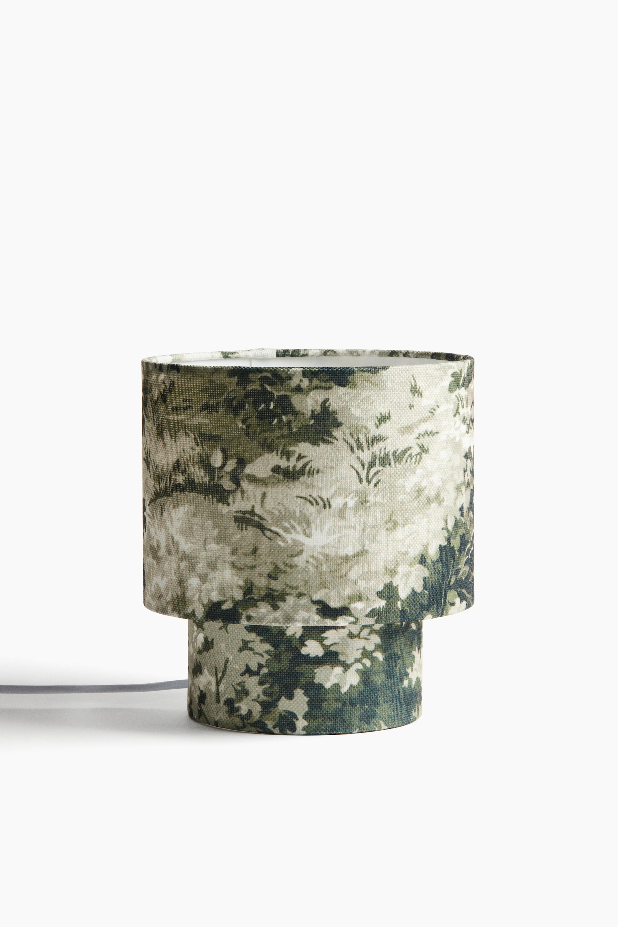 View larger image: A table lamp is facing front. It is featuring a two-tiered cylindrical shade and base, covered in a linen-look weave displaying an all-over print of green and gray foliage. A gray cord is extending from its base.