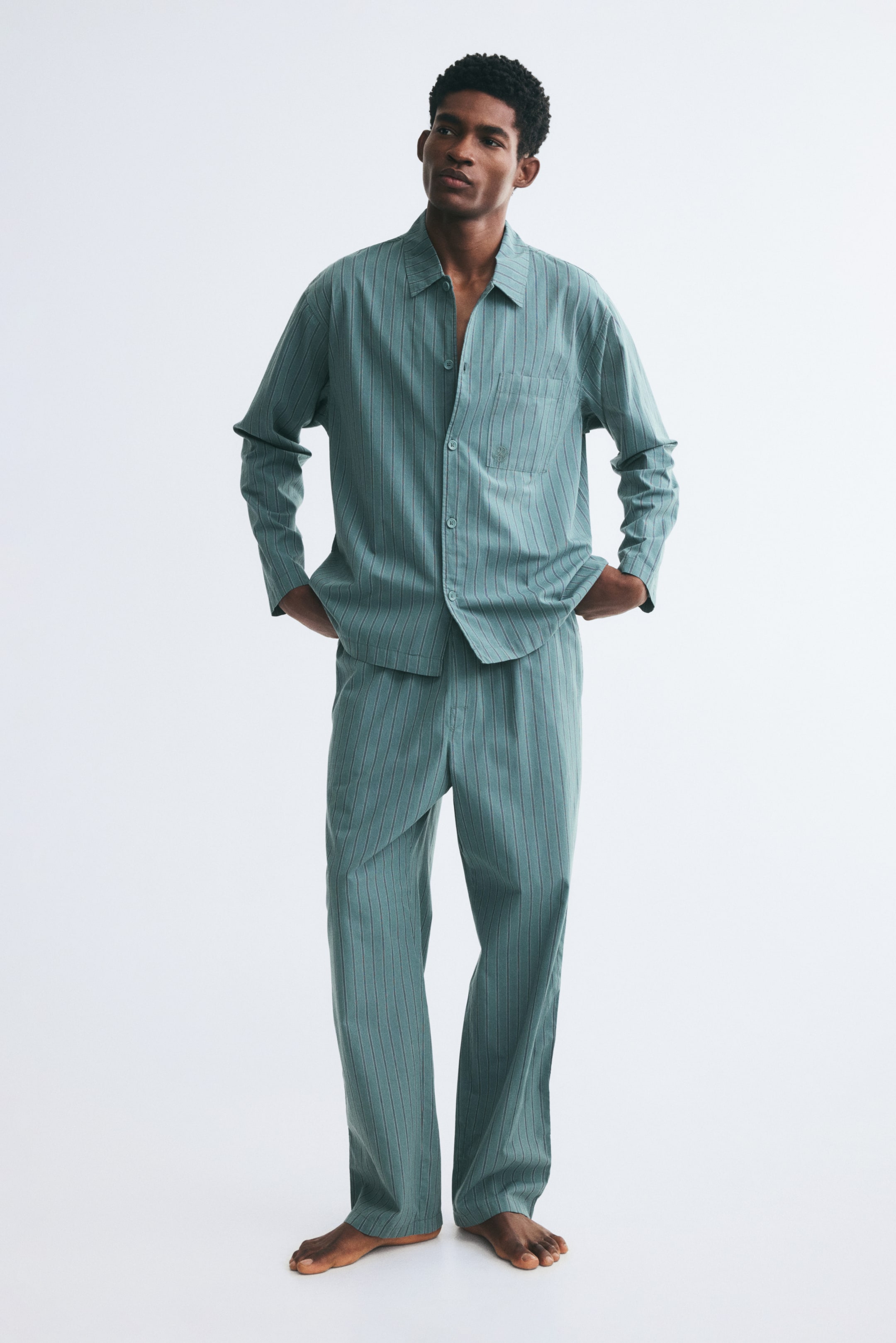 View larger image: The model is wearing a matching striped pyjama set in a muted green hue. The set features a long-sleeved, button-down shirt with a turn-down collar and a single chest pocket, paired with matching straight-leg pants.