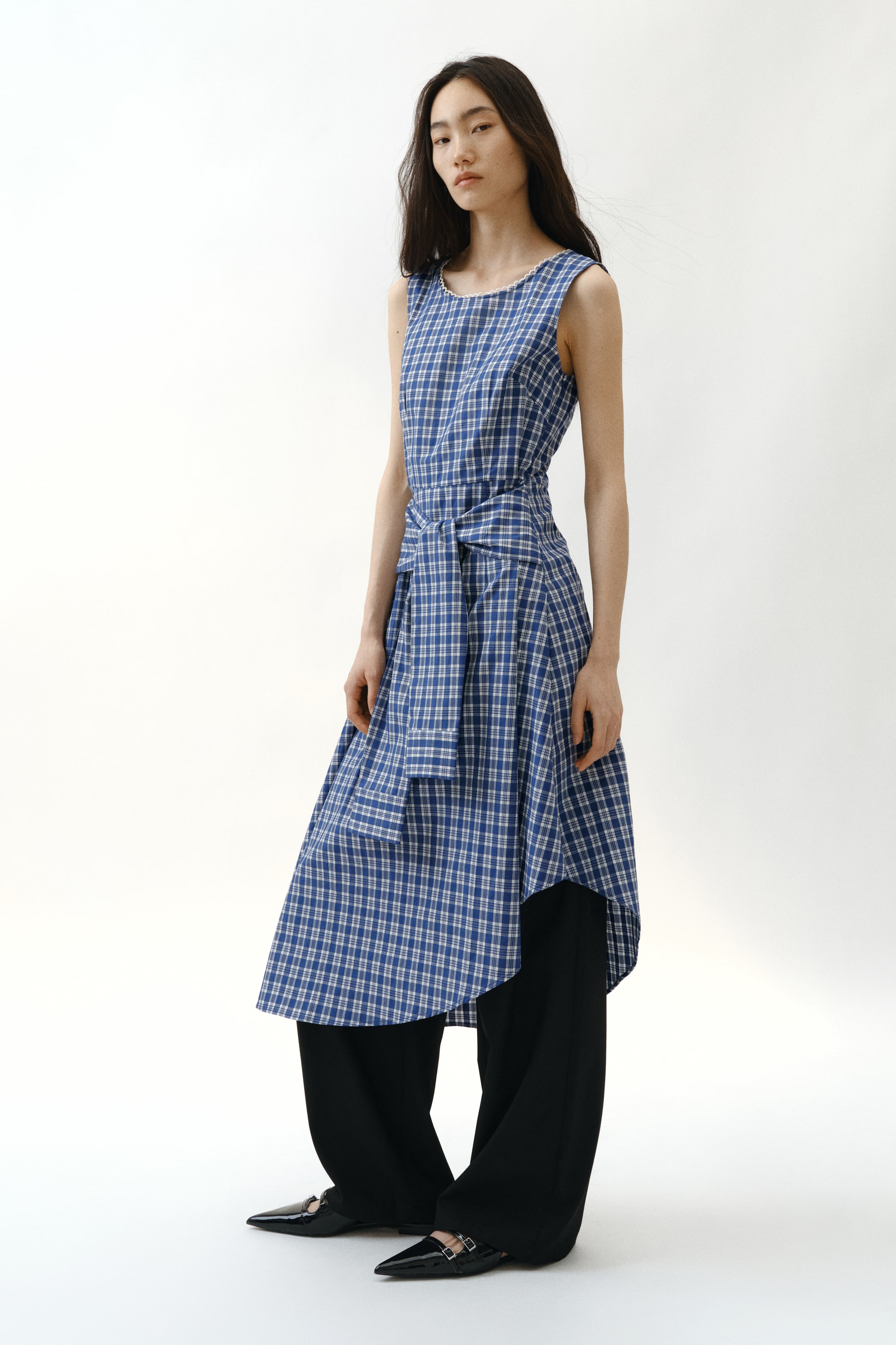 View larger image: A blue and white plaid sleeveless dress is showcased from a front-quarter angle, highlighting a round neckline with white trim and a front-tied waist. It features an asymmetrical hemline and is layered over black wide-leg pants.