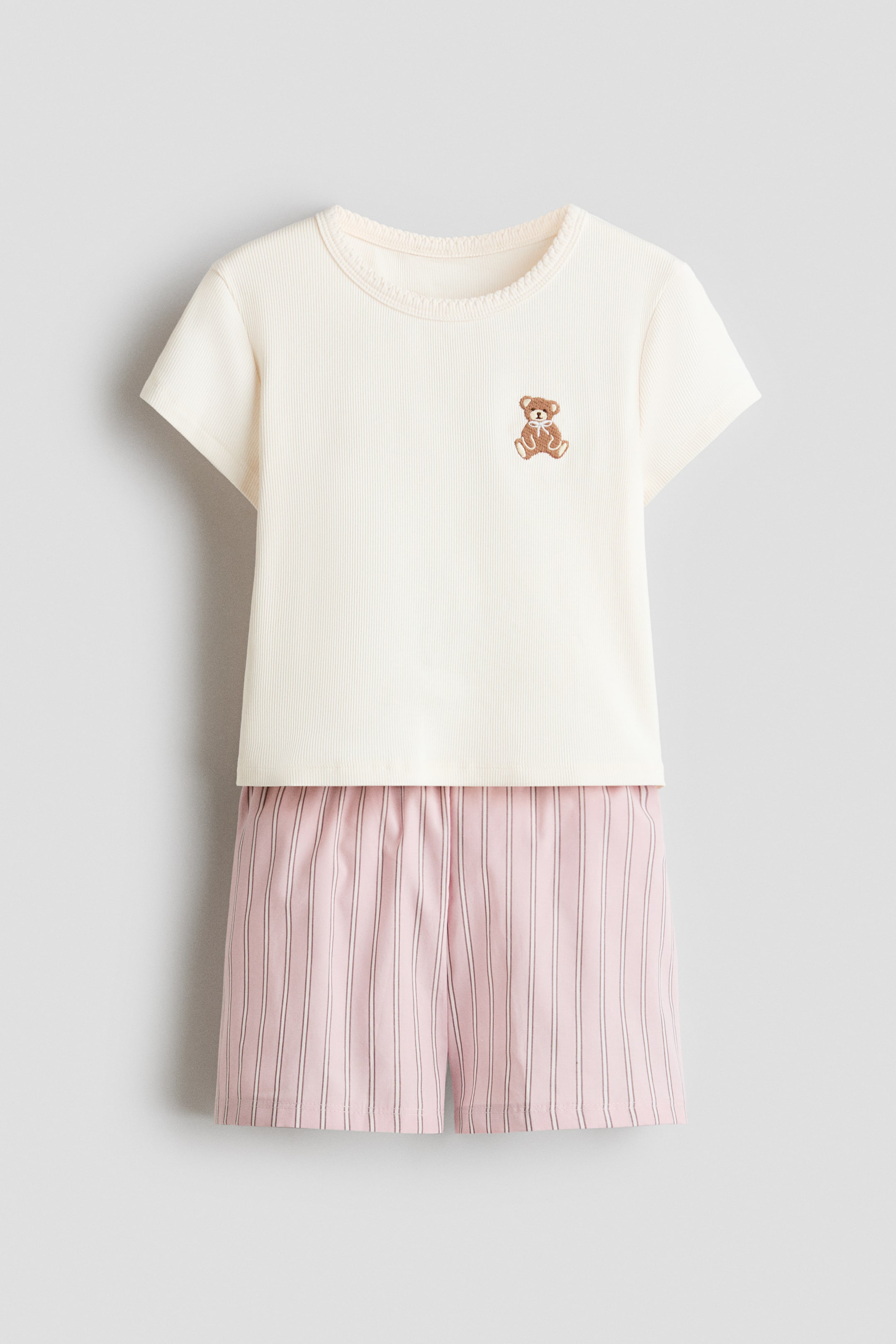 View larger image: A cream ribbed knit short-sleeved top with a scalloped trim along the crew neck and an embroidered brown teddy bear on the chest is paired with pink and white vertically striped shorts featuring an elasticated waistband, forming a sleepwear set.