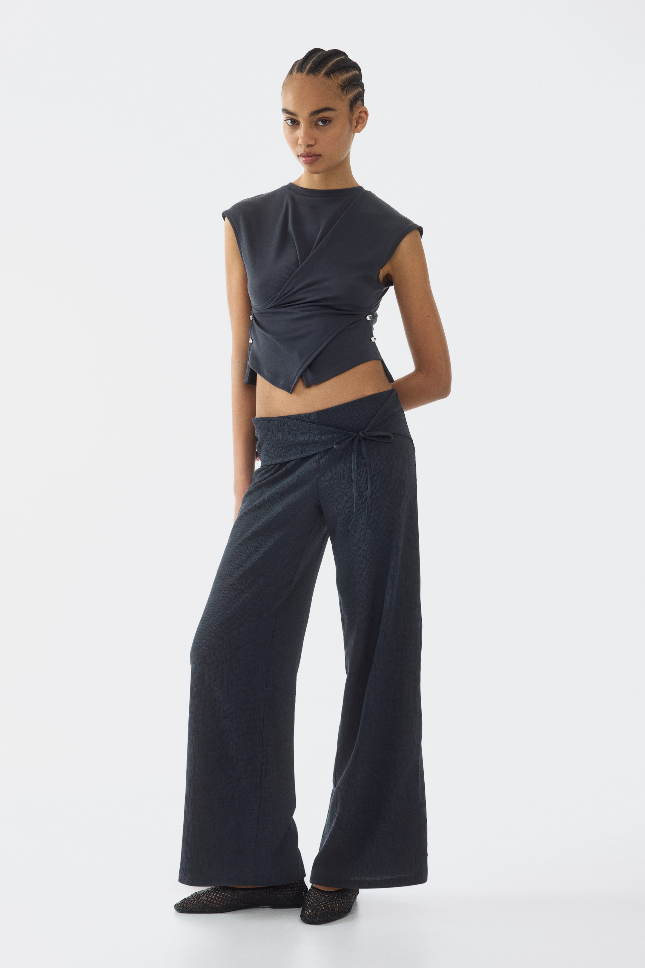 Büyük resmi görüntüle: Koyu gri bir takım, yapılı omuzları ve ön kısmı drapeli, gümüş tonlu zımbalarla sabitlenmiş kolsuz, kısa üst ile öne çıkıyor. Uyumlu geniş paça pantolonlar, ön kısmında bağlama detayı ve ince dikey çizgiler sergiliyor.