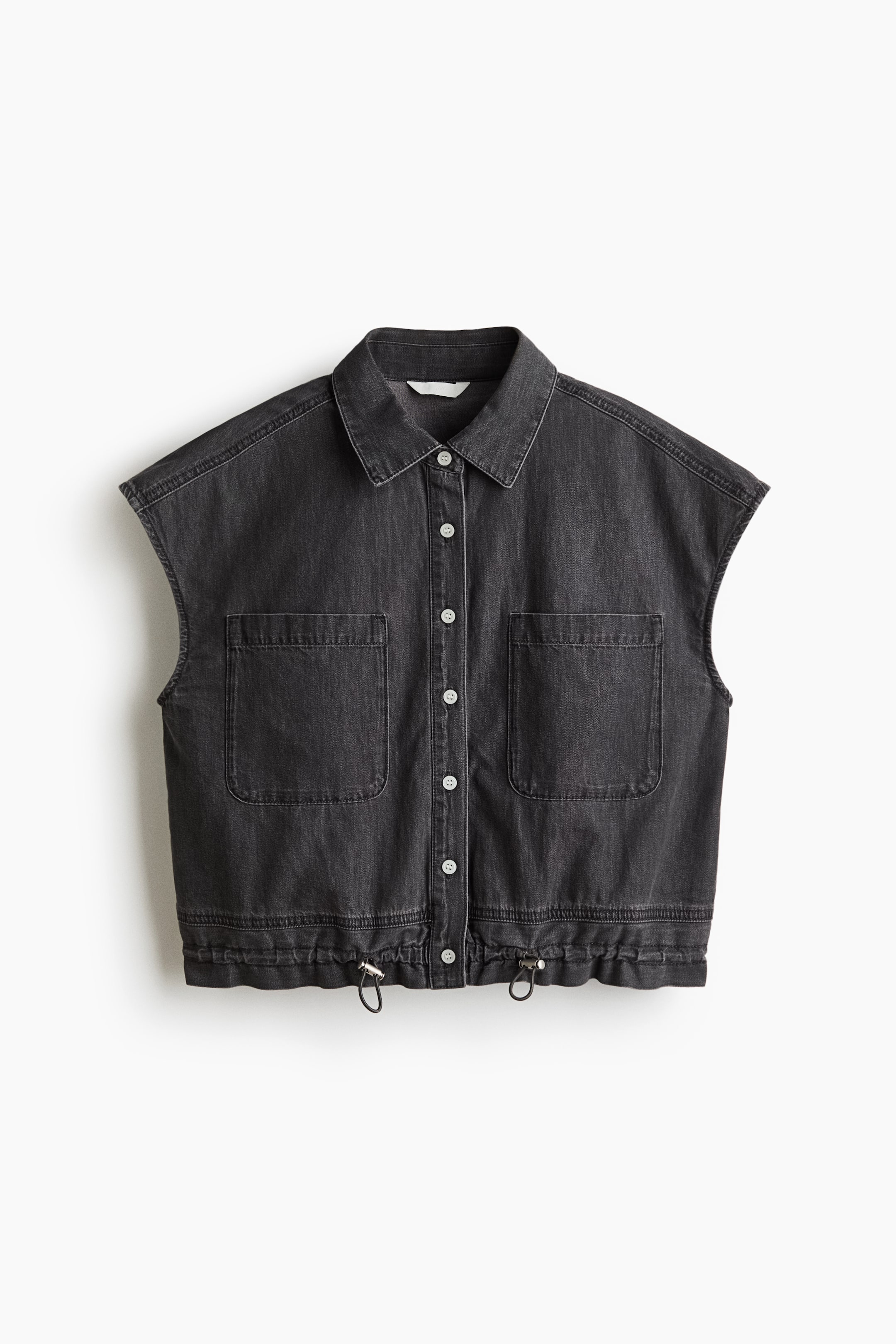 Xem ảnh lớn hơn: Một chiếc áo vest denim màu xám đậm đang hướng mặt trước, nổi bật với cổ áo nhọn cổ điển và nút cài phía trước với các nút màu trắng. Hai túi lớn dạng miếng vá xuất hiện trên ngực, và gấu áo được thiết kế với dây rút có thể điều chỉnh và các nút bạc.