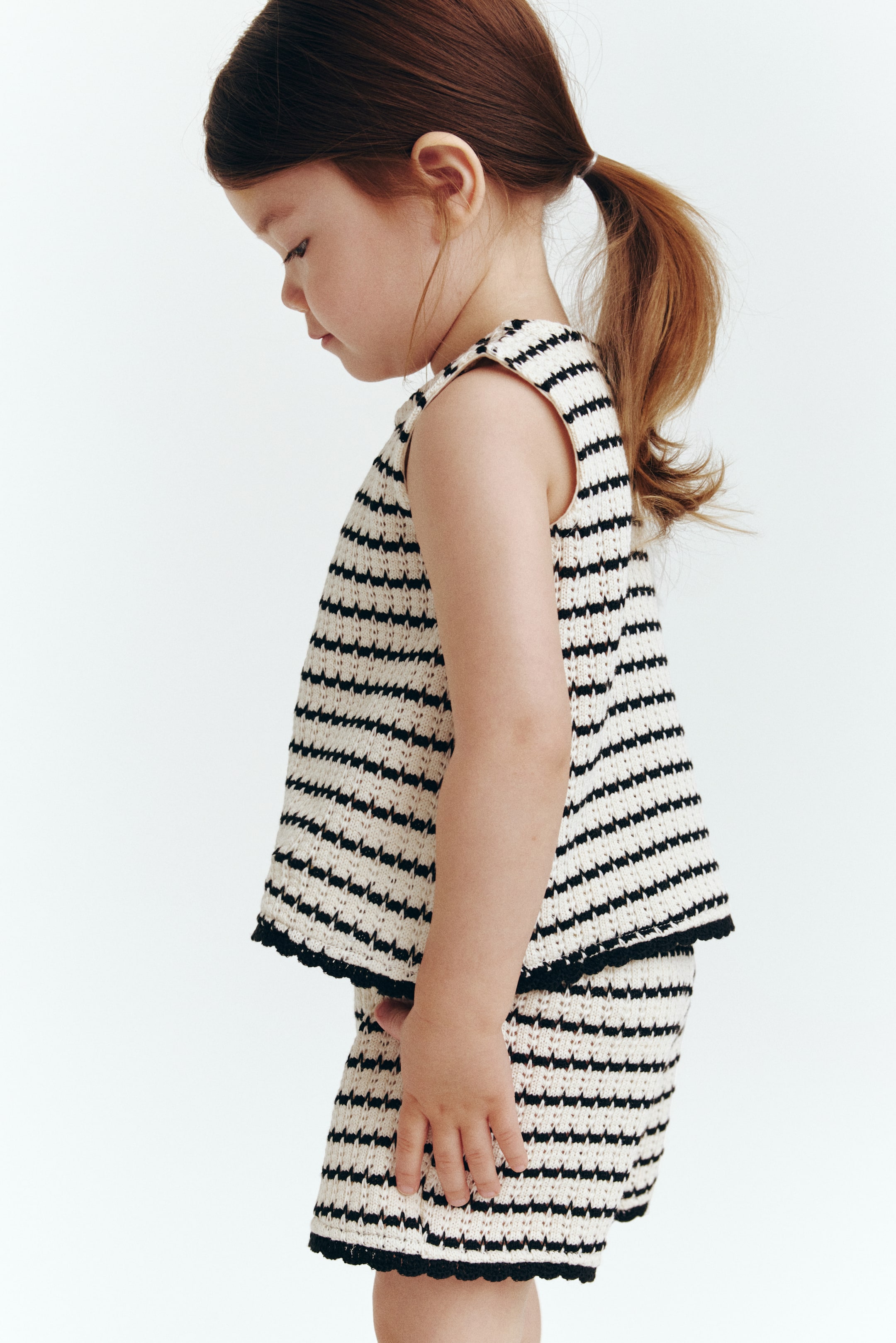 Agrandir l'image: Un ensemble en tricot deux pièces pour enfant, avec des rayures texturées horizontales crème et noires, est présenté de côté. Le haut sans manches et le short assorti mettent en valeur une bordure festonnée sombre le long de leurs ourlets.