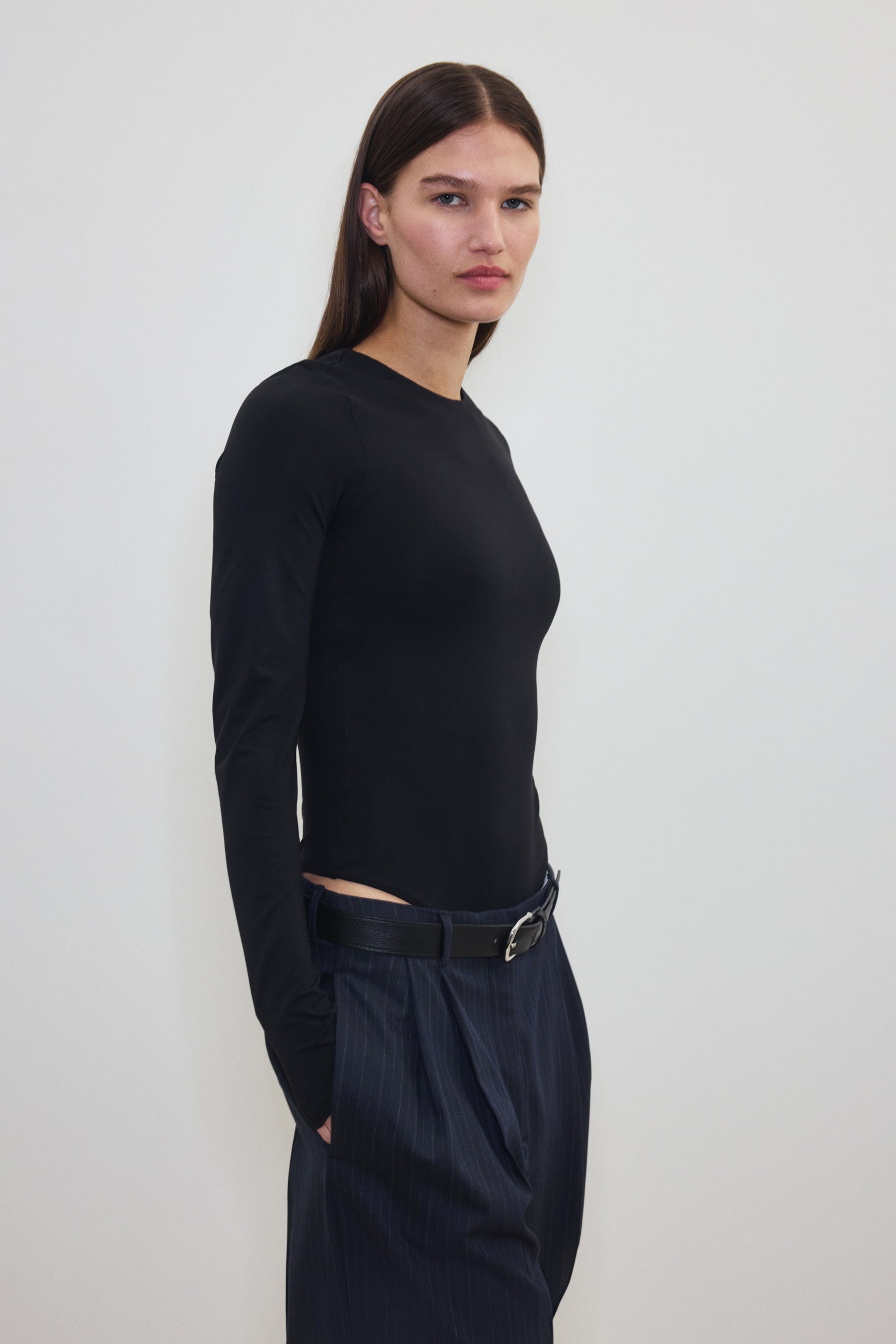 Größeres Bild ansehen: Ein schwarzer langärmeliger Body mit rundem Ausschnitt wird von dem Model getragen. Der Body sitzt glatt und ist in dunkle Nadelstreifenhosen gesteckt, die von einem schwarzen Gürtel gehalten werden.