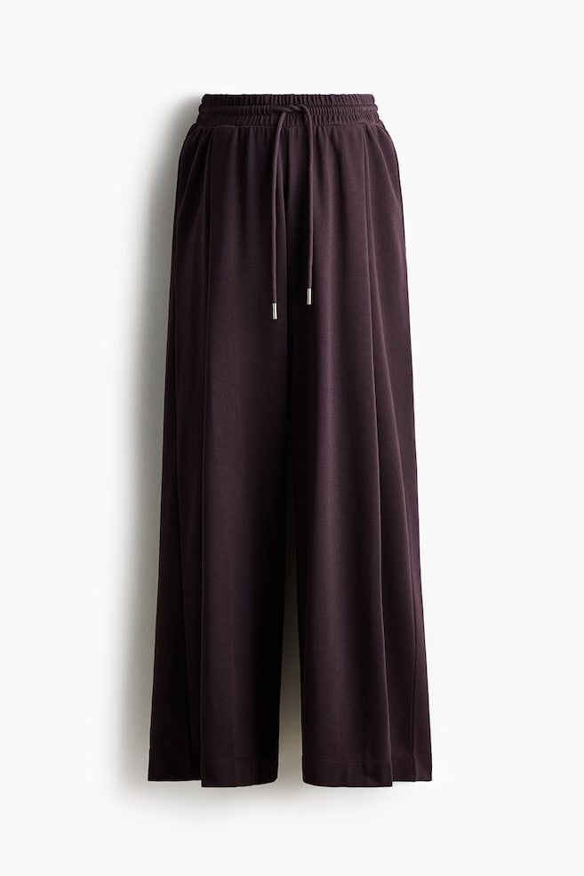 A pair of deep plum wide-leg trousers is shown from the front, featuring an elasticated waistband with a matching drawstring finished with silver-tone metal aglets. The trousers drape downward in a loose, relaxed fit. - 1