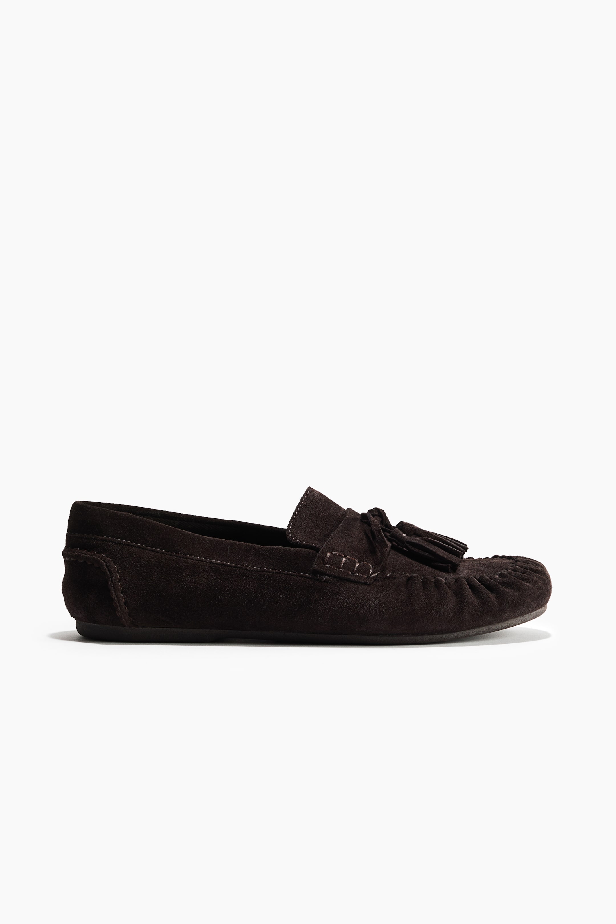 View larger image: A dark brown suede loafer showcases its side profile, highlighting prominent tassels on the vamp and a moc-toe construction with visible stitching. A flat dark sole supports the shoe.