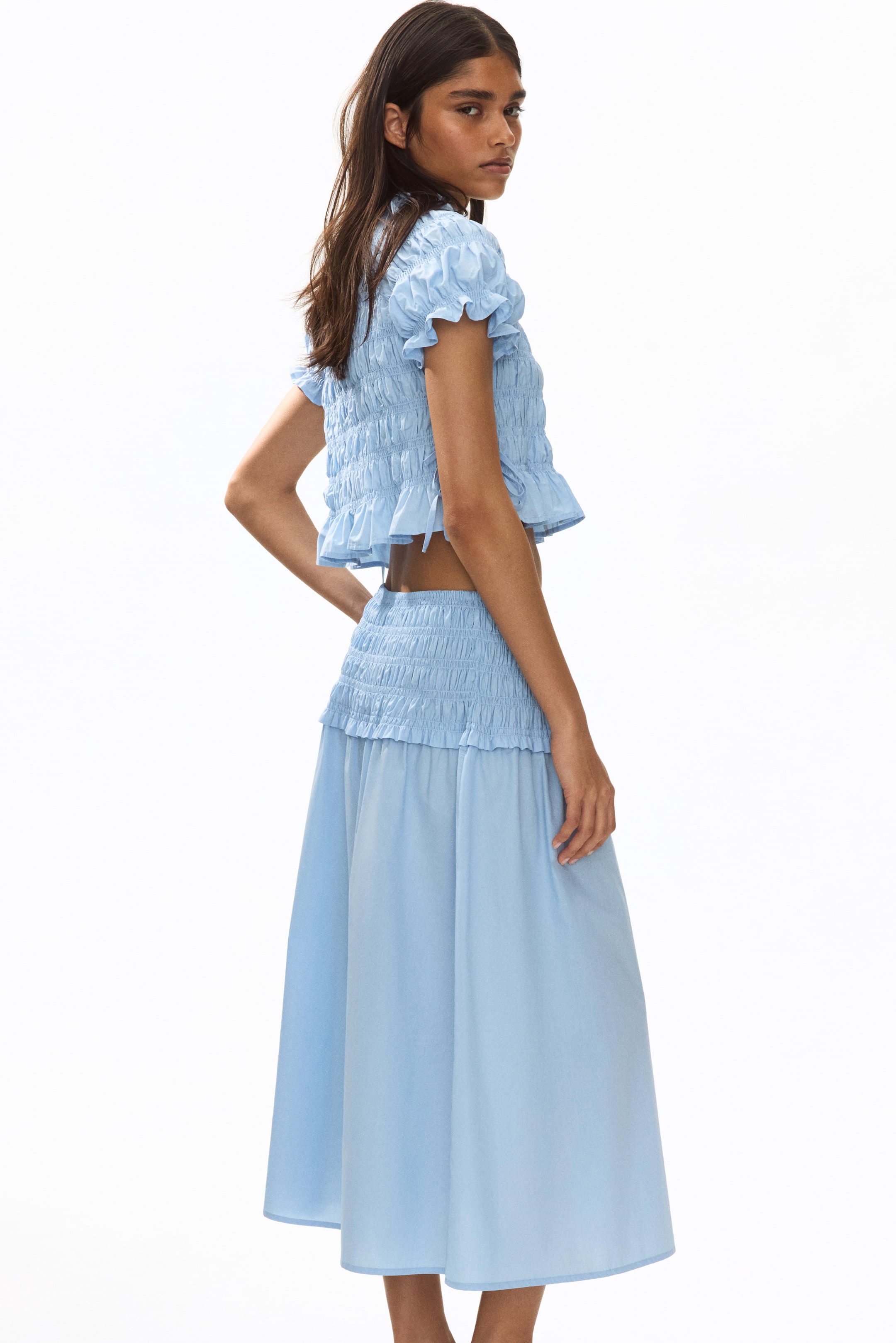 View larger image: A light blue two-piece set is showcased from a side-back angle, highlighting a cropped top with a smocked bodice, short puffed sleeves with ruffled cuffs, and a ruffled hem with side ties, perfectly paired with a midi skirt featuring a smocked waistband and a flowing silhouette.