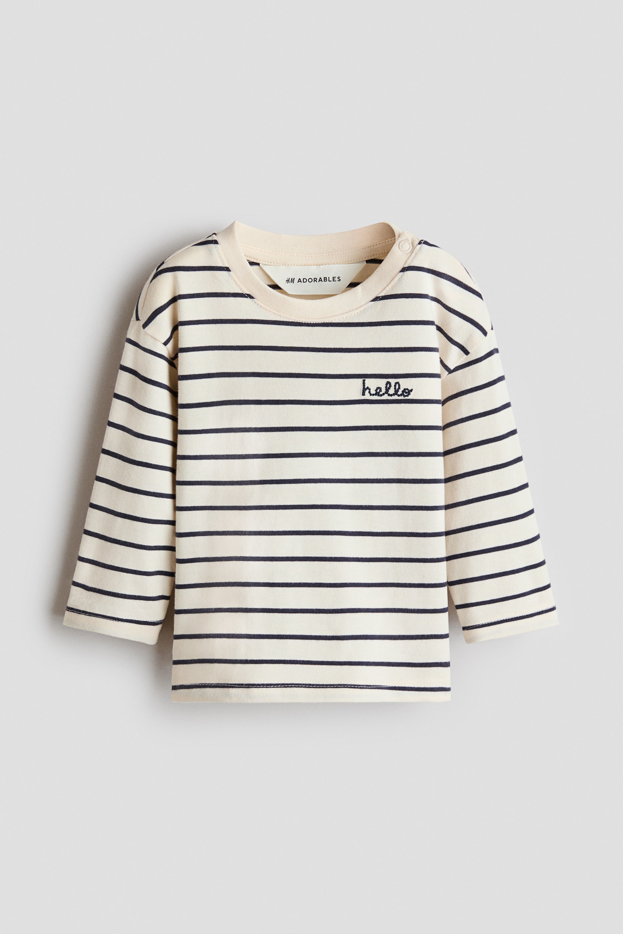 View larger image: A loose-fit T-shirt is shown from the front, featuring cream and navy horizontal stripes. It is designed with a rib-trimmed round neckline, with press studs visible on one side. A small "hello" motif is embroidered on the upper left front, and long sleeves with dropped shoulders are visible.