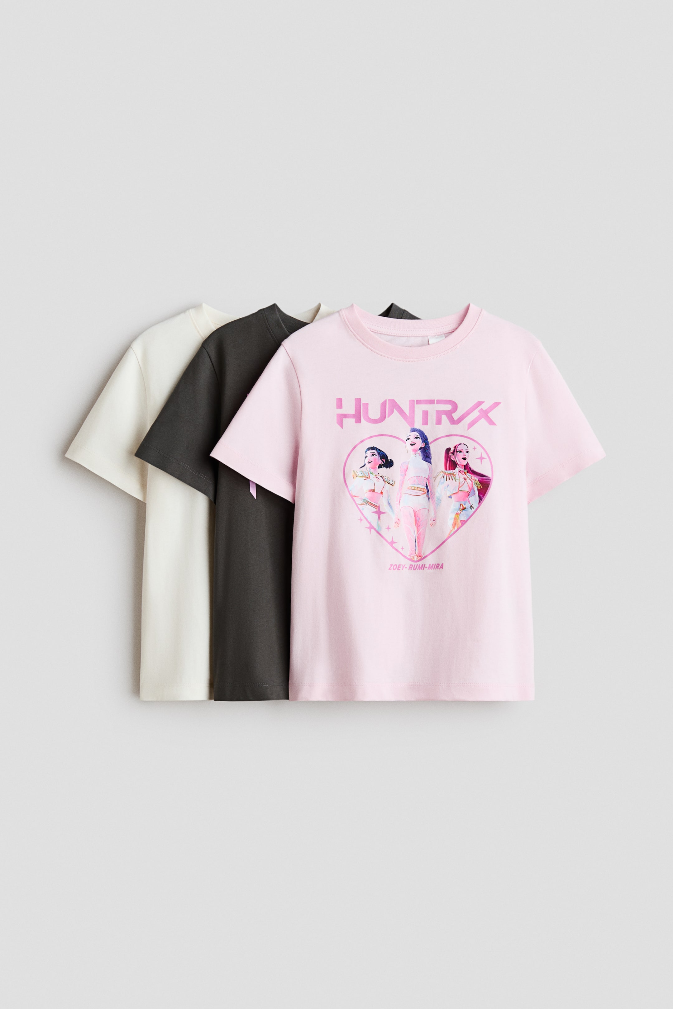 Größeres Bild ansehen: Drei kurzärmelige Oberteile aus weichem Baumwolljersey sind übereinander geschichtet, von vorne nach hinten in Pink, Dunkelgrau und Off-White präsentiert. Das vordere pinke Oberteil zeigt ein Druckmotiv mit dem Text „HUNTRX“ und drei stilisierten weiblichen Figuren innerhalb einer Herzumrandung.
