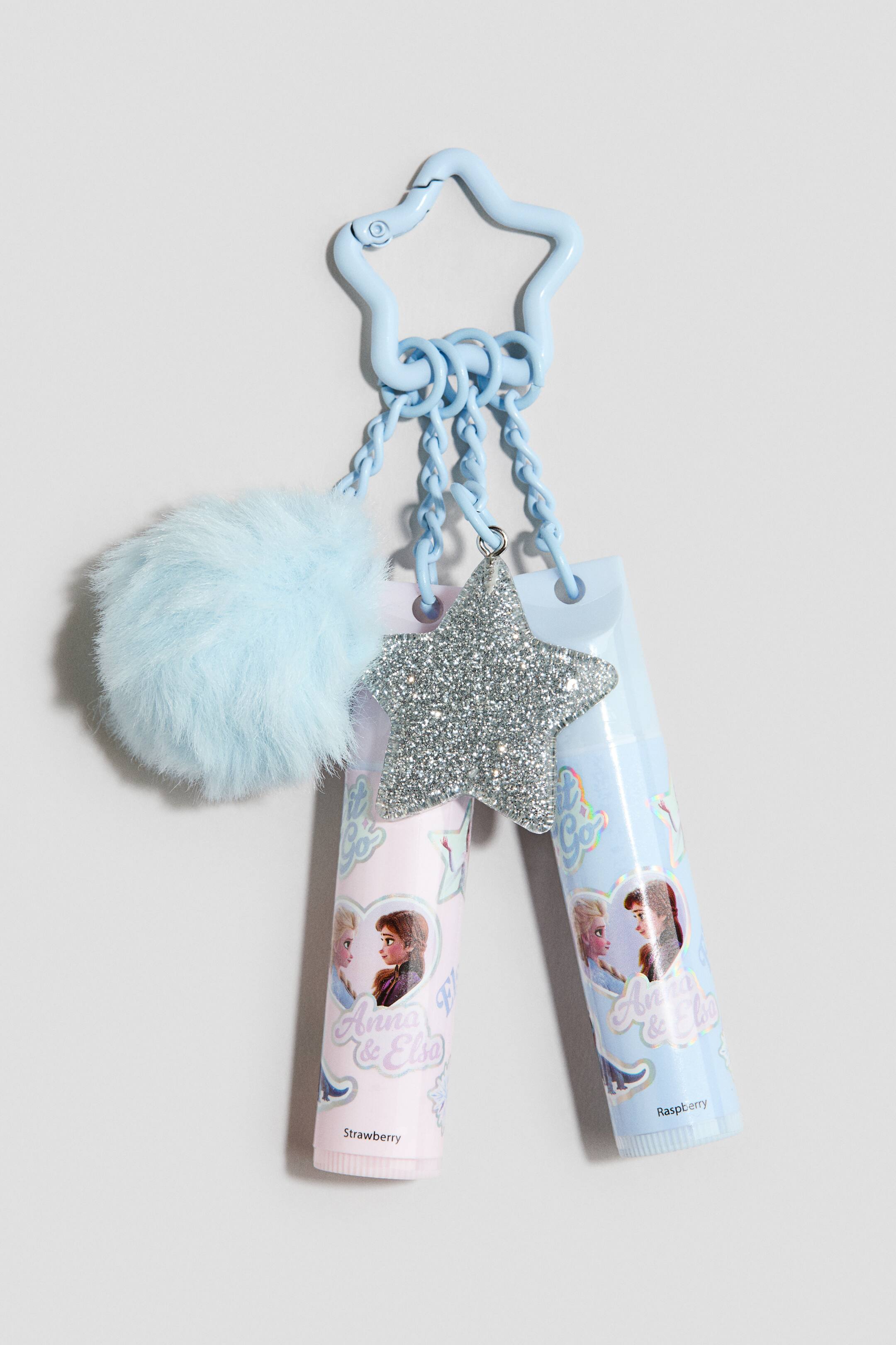 View larger image: A light blue star-shaped keyring clasp holds three dangling chains featuring a fluffy light blue pom-pom, a silver glittery star charm, and two lip balm tubes. The pink and light blue lip balm tubes are printed with character illustrations and holographic text, displaying the flavours "Strawberry" and "Raspberry" at the bottom.