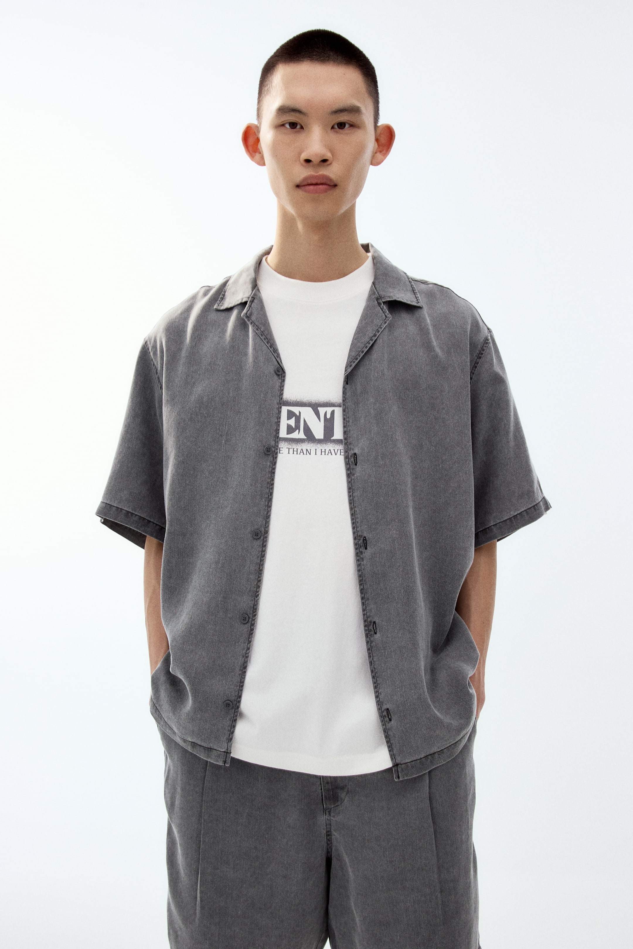 View larger image: A washed grey short-sleeved shirt, featuring a relaxed Cuban collar and a button-front placket, is worn open over a white graphic T-shirt. The shirt is paired with matching relaxed grey shorts, shown from a front view.