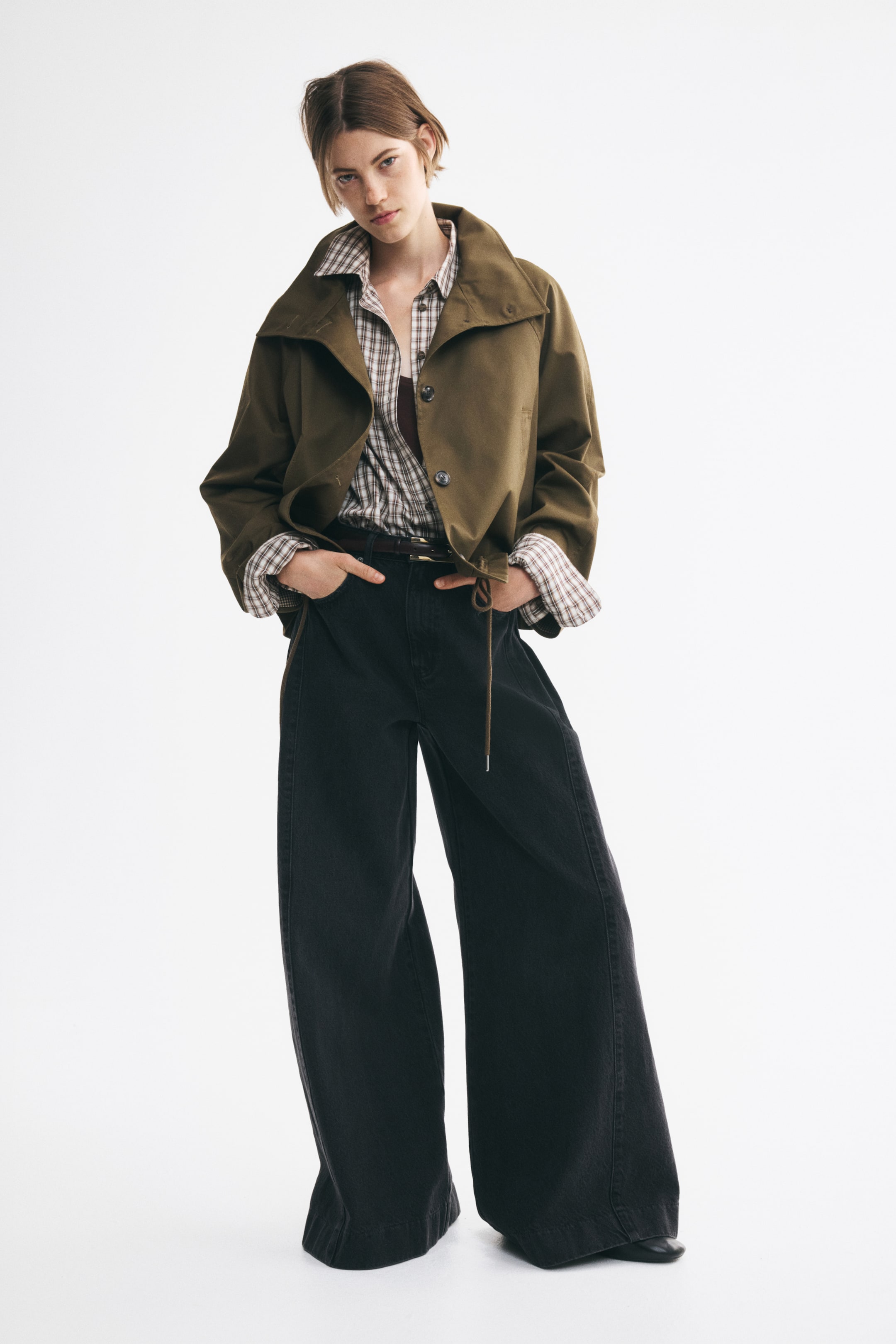 Büyük resmi görüntüle: Önden görünümde, yanlarda ters kutu pili detaylarıyla zenginleştirilmiş, bol kesimli ve ekstra geniş paçalara sahip siyah denim pantolon giyen bir model yer alıyor. Normal bel kesimli pantolon, kemerle kombinlenmiş olup ayak hizasında minimal yığılmayla sonlanarak yapılandırılmış ve hacimli siluetini öne çıkarıyor.