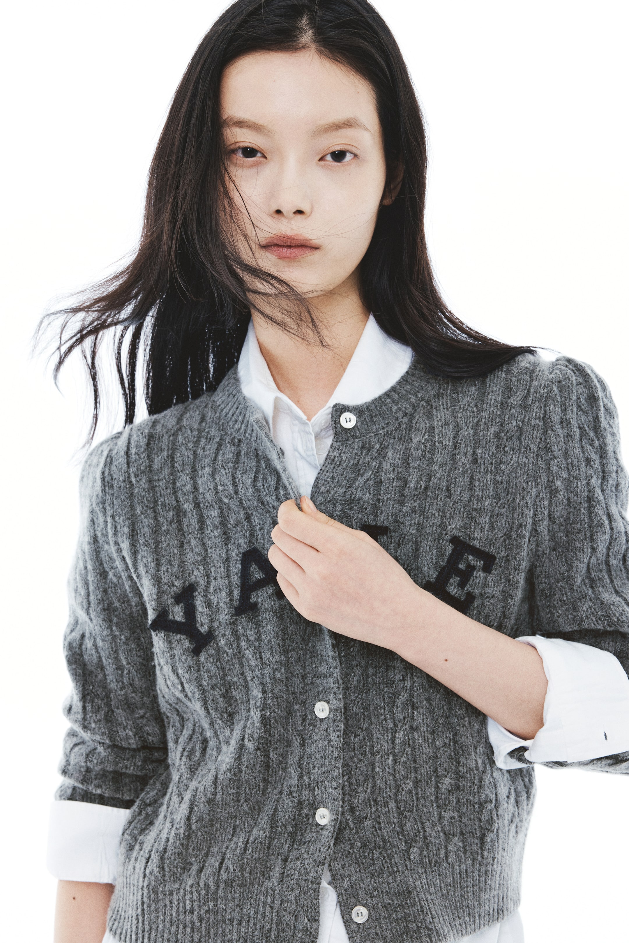 이미지 확대 보기: 회색의 텍스처가 있는 니트 카디건으로, 흰색 버튼과 가슴 부분의 검은색 컬리지 레터링이 특징이며, 모델은 흰색 칼라 셔츠 위에 착용하고 있습니다. 모델은 한 손으로 칼라 근처에서 카디건 앞여밈을 잡고 있습니다.
