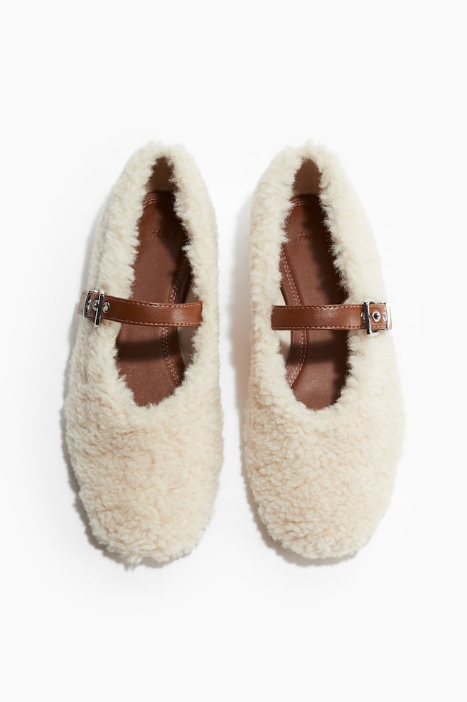 A pair of Mary Jane-style ballet pumps is being displayed facing front, featuring a fluffy, cream-colored fabric with almond toes. Each shoe is showcasing an adjustable brown patent-finish foot strap with a silver-toned metal buckle and a visible brown jersey lining. - 1