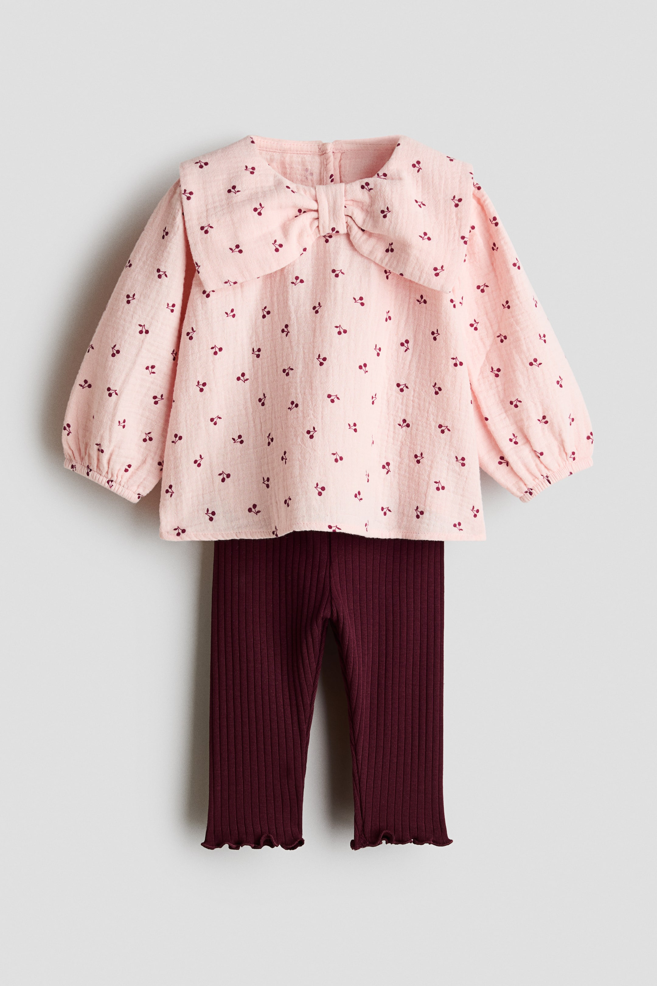 View larger image: This set showcases a light pink muslin blouse, presented from the front, adorned with a cherry pattern, featuring an oversized bow-shaped collar and elasticated cuffs on its long sleeves. It is paired with dark red ribbed jersey leggings, which exhibit a ribbed texture and wavy overlocked hems.