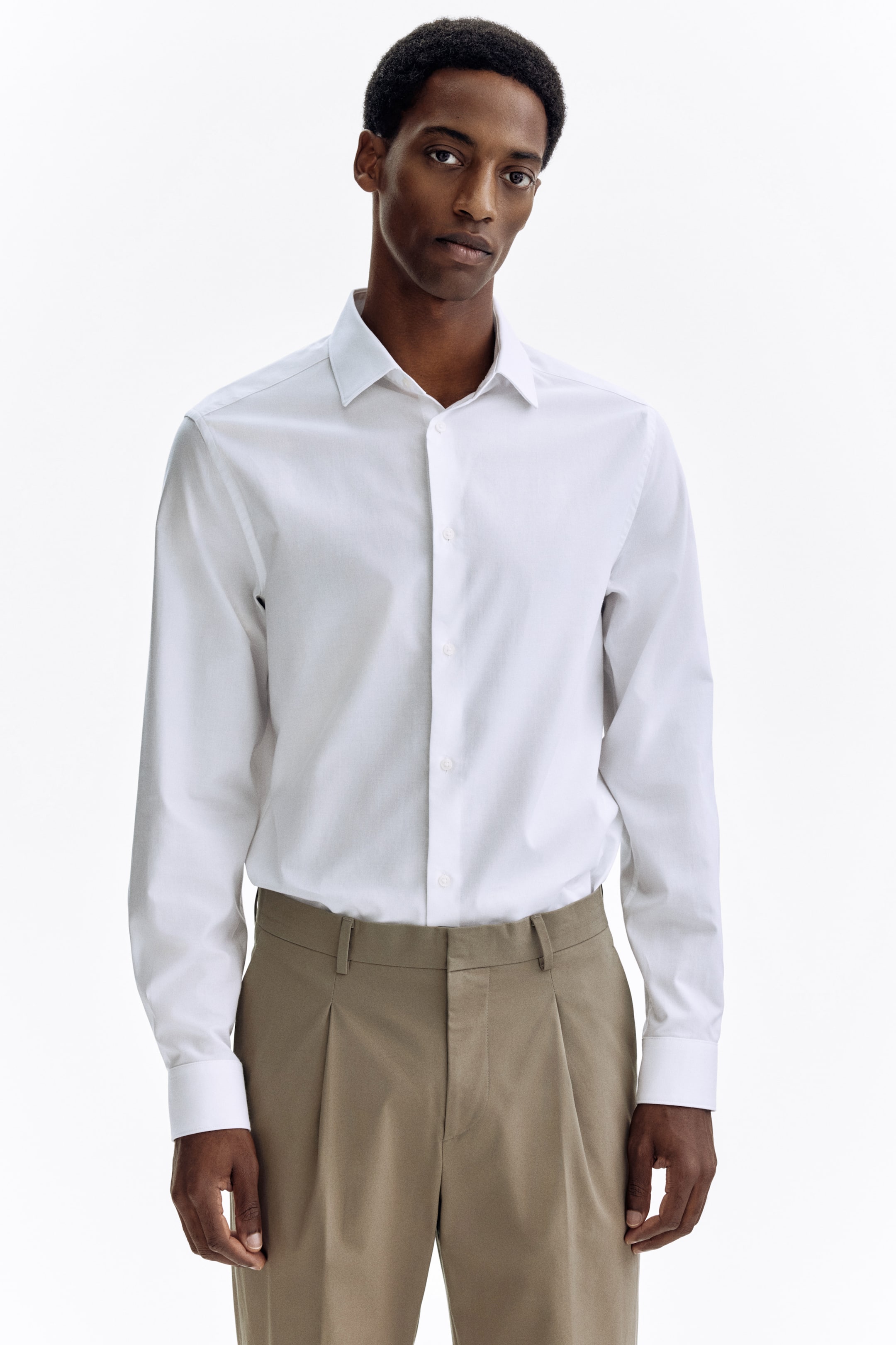 Agrandir l'image: La chemise blanche à boutons est dotée d'un col en pointe, de manches longues avec poignets boutonnés, et d'une patte de boutonnage visible avec des boutons blancs. Elle est rentrée dans un pantalon plissé marron clair.