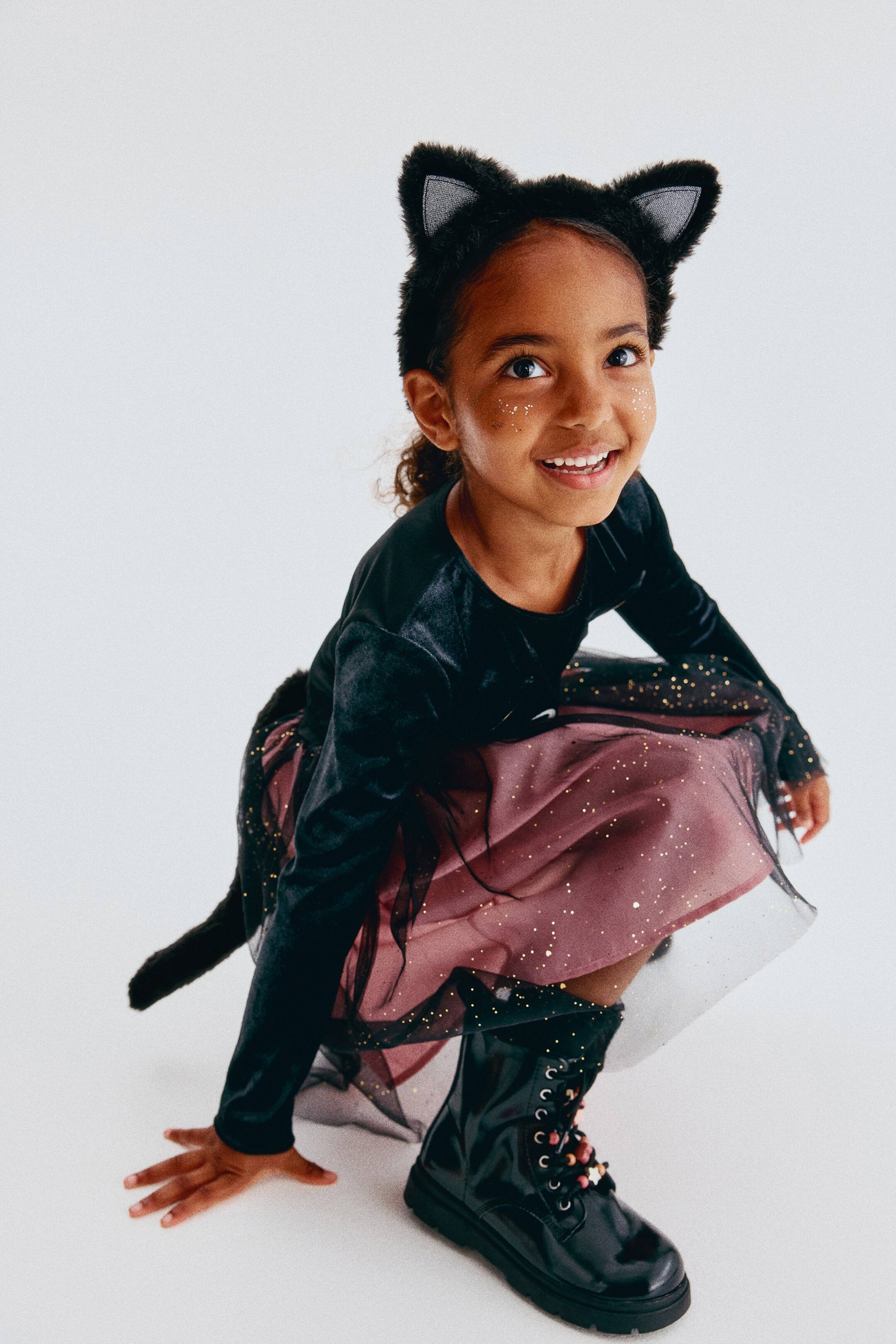 Grotere afbeelding bekijken: Deze tweedelige katten-fancy dressset wordt getoond aan een kind dat gehurkt en naar voren gericht staat, met een jurk met lange mouwen, een zwart fluwelen lijfje, een roze rok met een zwarte tule overlay met goudspikkels en een vastgemaakte zwarte kattenstaart. De set wordt gecompleteerd door een bijpassende zachte zwarte haarband met puntige kattenoren met zilverkleurige glinsterende binnenpanelen.