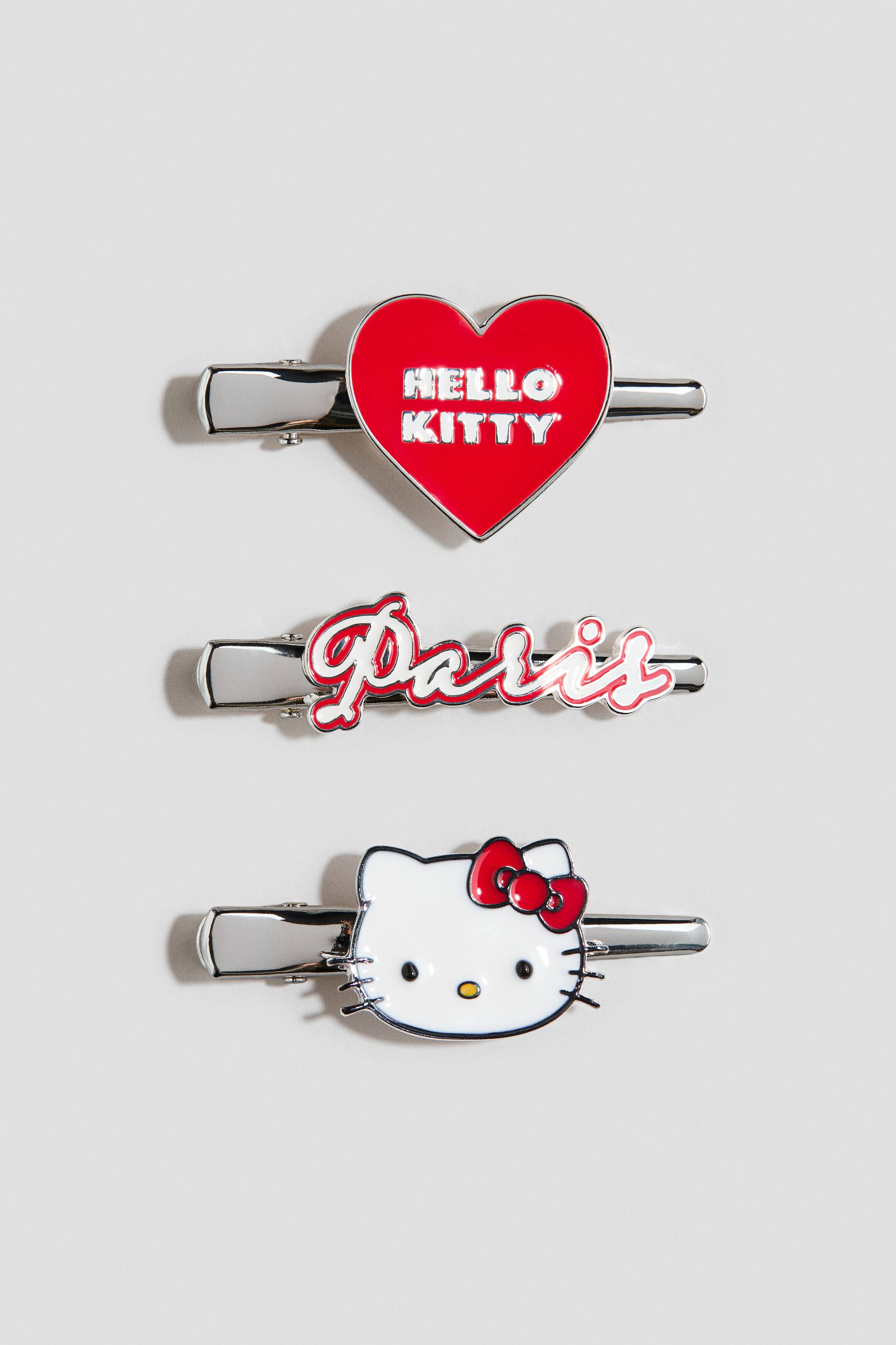 Ampliar la imagen: Tres pinzas para el cabello están mostrando diseños de esmalte sobre bases de pinza de presión de color plateado. La pinza superior muestra un corazón rojo con "HELLO KITTY" en letras plateadas, la pinza del medio presenta "Paris" en escritura roja con un contorno blanco, y la pinza inferior muestra una cabeza blanca de Hello Kitty con un lazo rojo.