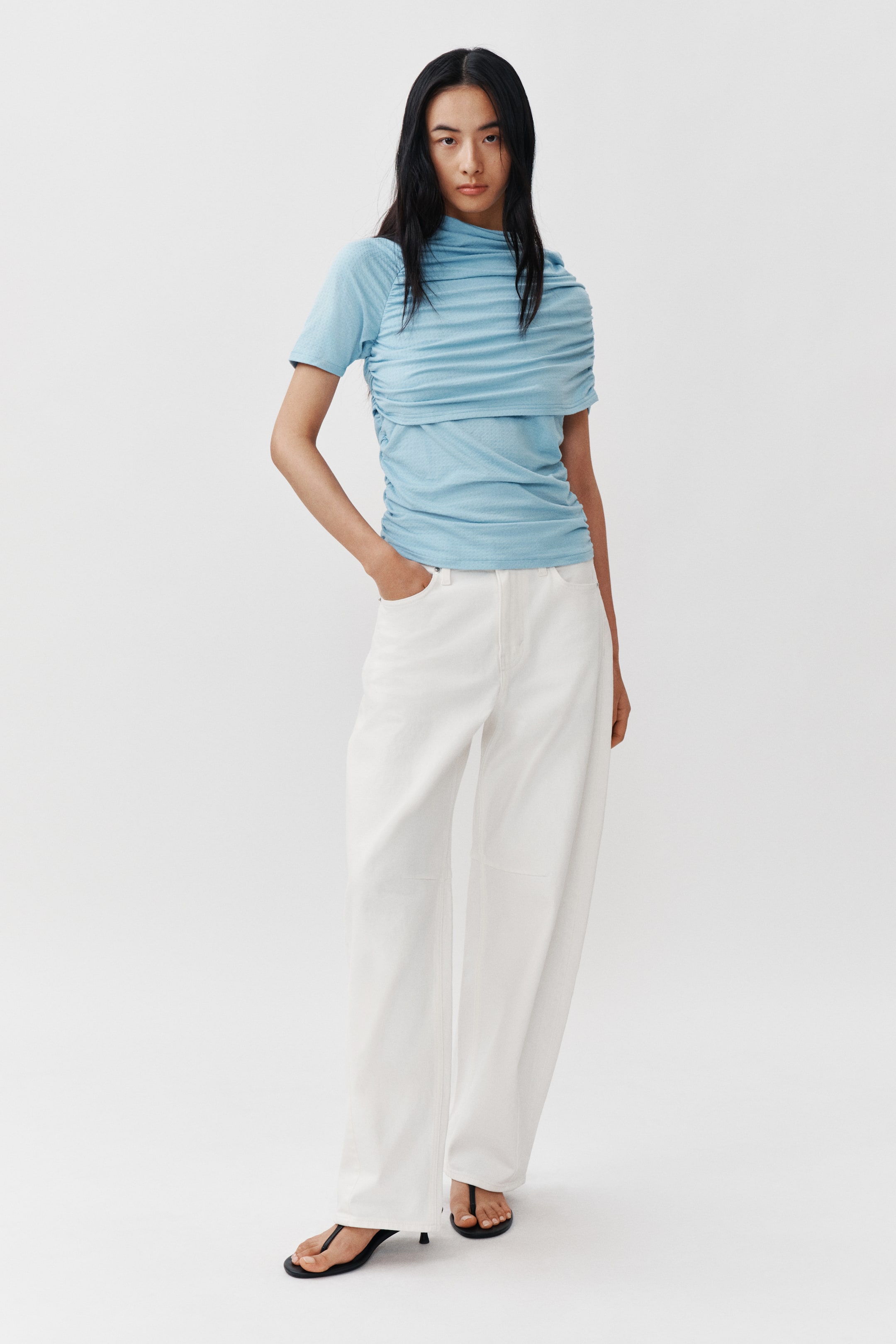 View larger image: A light blue short-sleeved top is featuring extensive horizontal ruching across the chest and body, with the fabric forming a high, draped neckline. It is paired with white, high-waisted, wide-leg trousers.