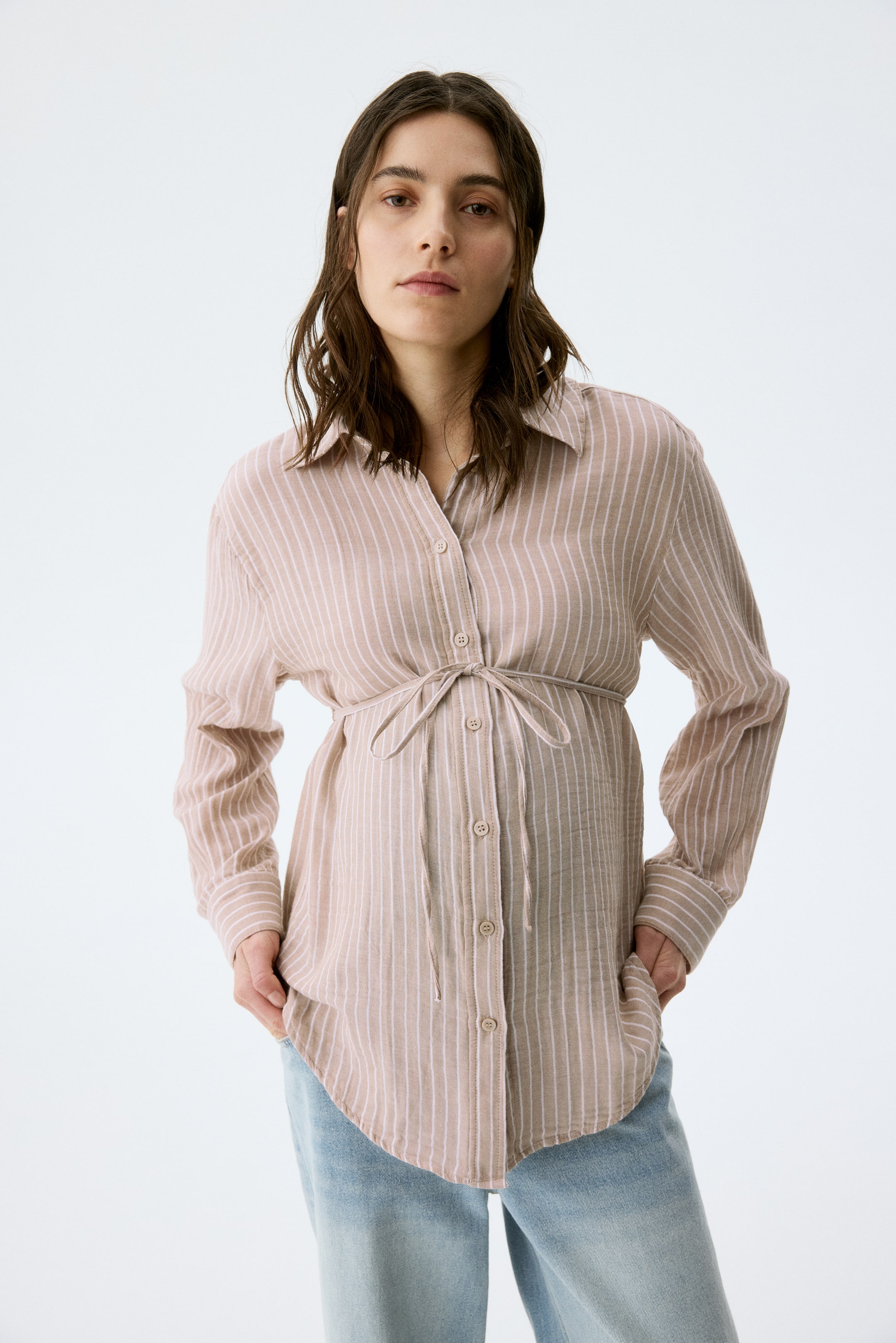 View larger image: "Isang beige na button-down na kamiseta na may manipis na puting patayong guhit ay may pointed na kwelyo, button-down na harapan, at mahahabang manggas na may buttoned cuffs. Isang self-fabric na sinturon ang nagtatali sa baywang, na lumilikha ng gathered na detalye, at ang kamiseta ay may rounded na laylayan."