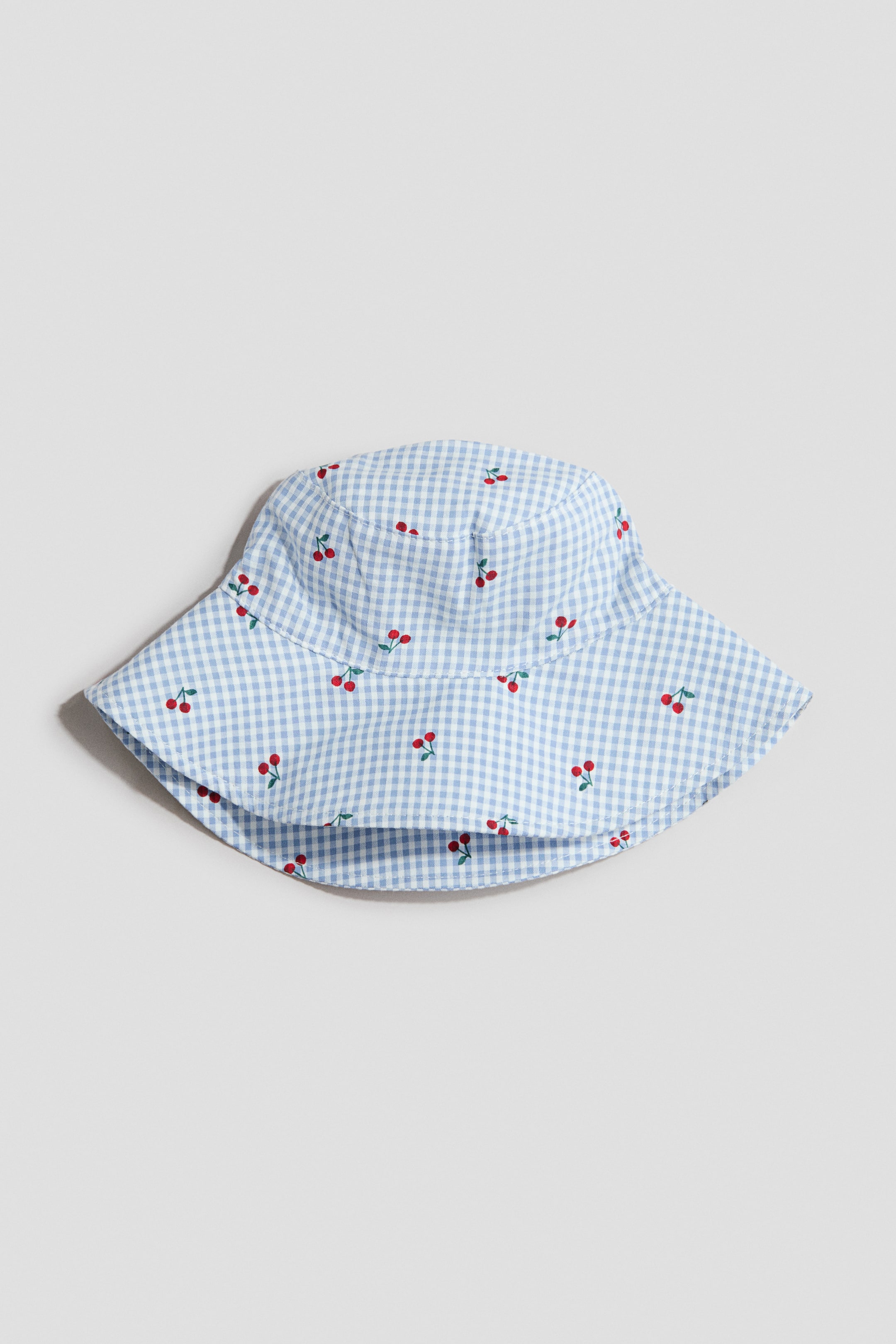 Grotere afbeelding bekijken: Een zonnehoed met een lichtblauw en wit gingham patroon, versierd met verspreide rode kersenmotieven. Het ontwerp heeft een afgeronde kroon en een brede rand, gepresenteerd vanaf de voorkant.