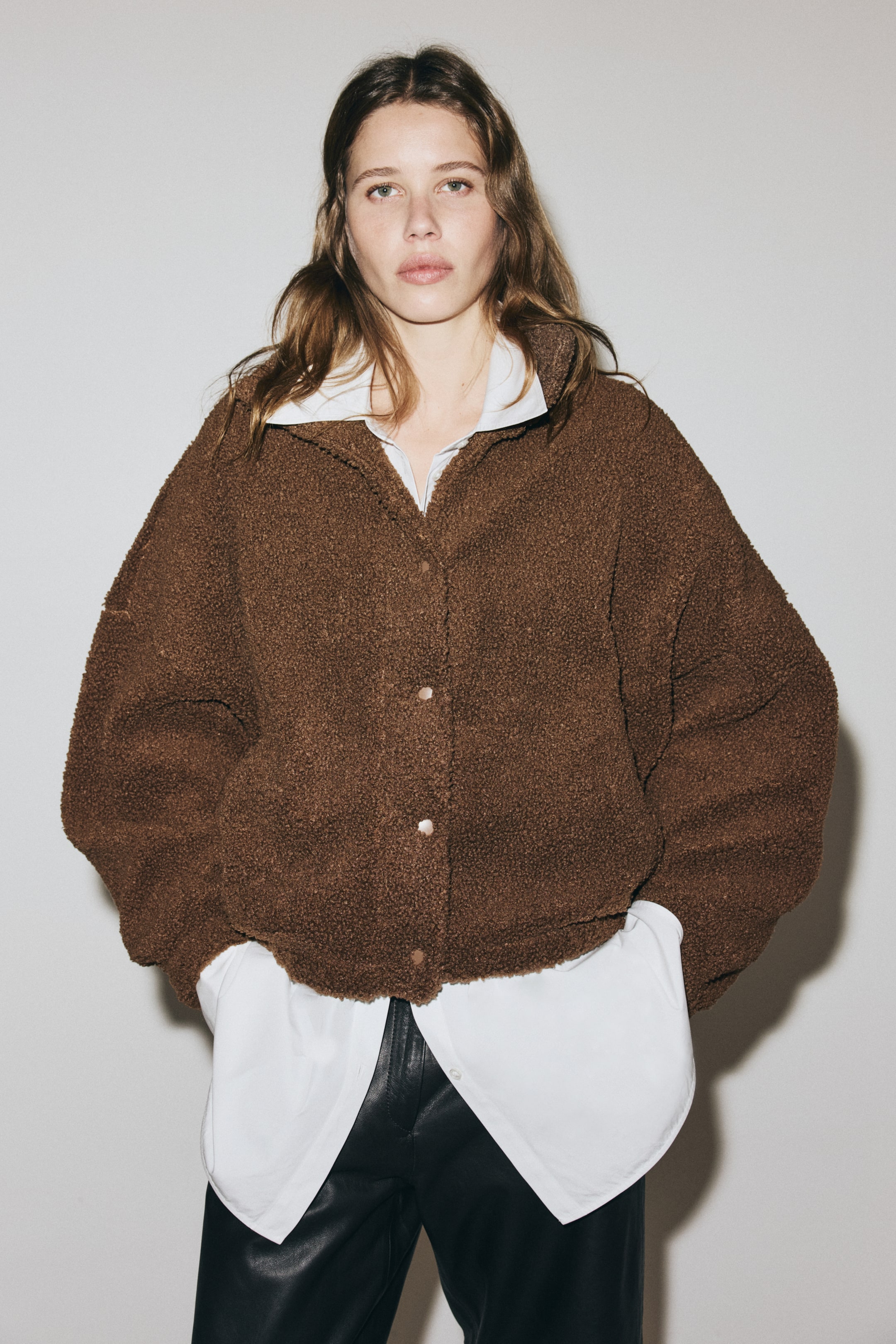 Vis større bilde: En brun teddijakke presenteres fra forsiden, stylet over en hvit skjorte. Den har en høy krage, knappelukning foran, lav skuldersøm og skrålommer på sidene.