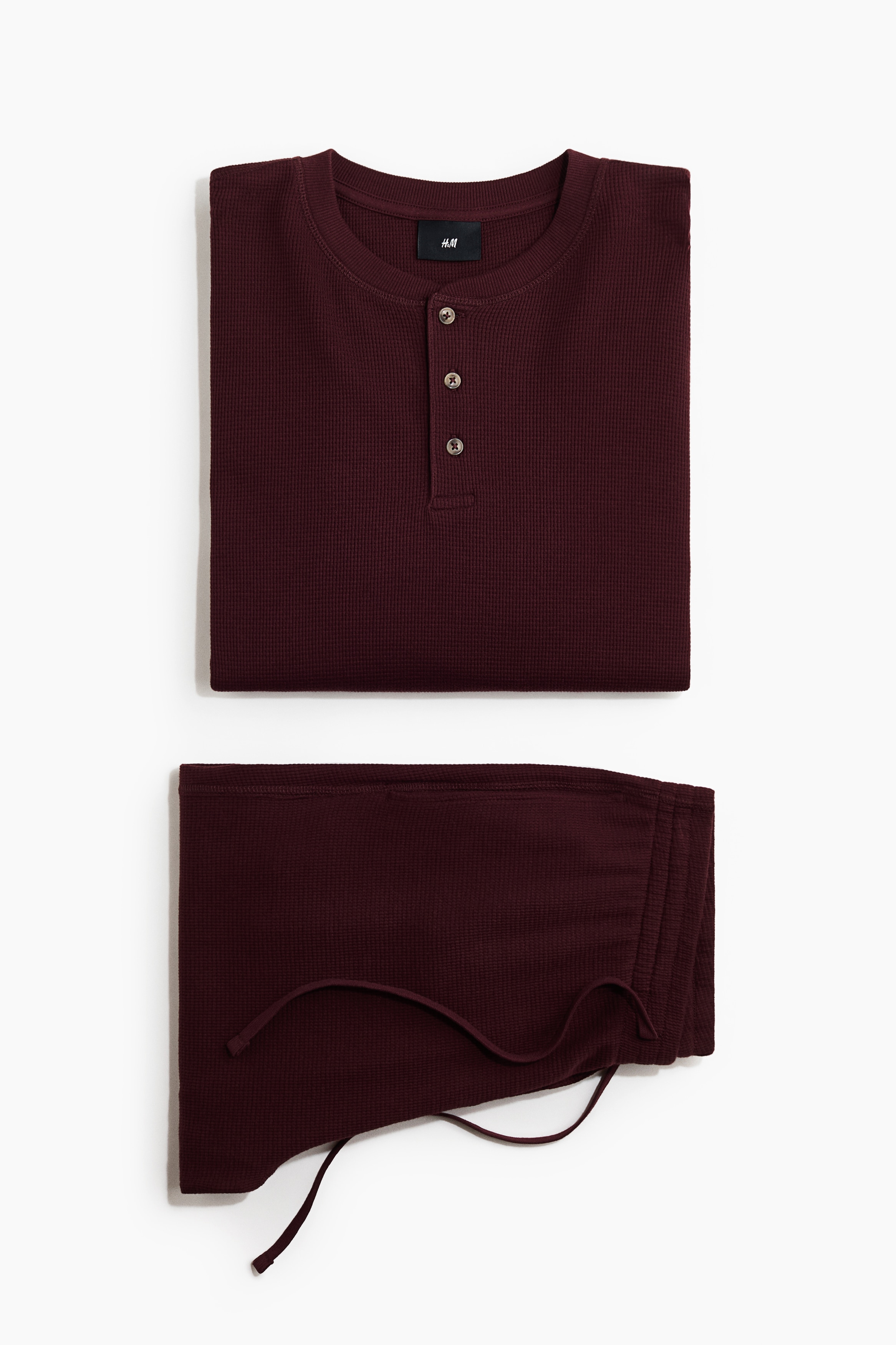 Das Model trÃ¤gt ein bordeauxfarbenes Loungewear-Set aus geripptem Baumwolljersey. Das Set umfasst ein Langarmshirt mit Knopfleiste und eine passende gerade Hose mit elastischem Tunnelzugbund.