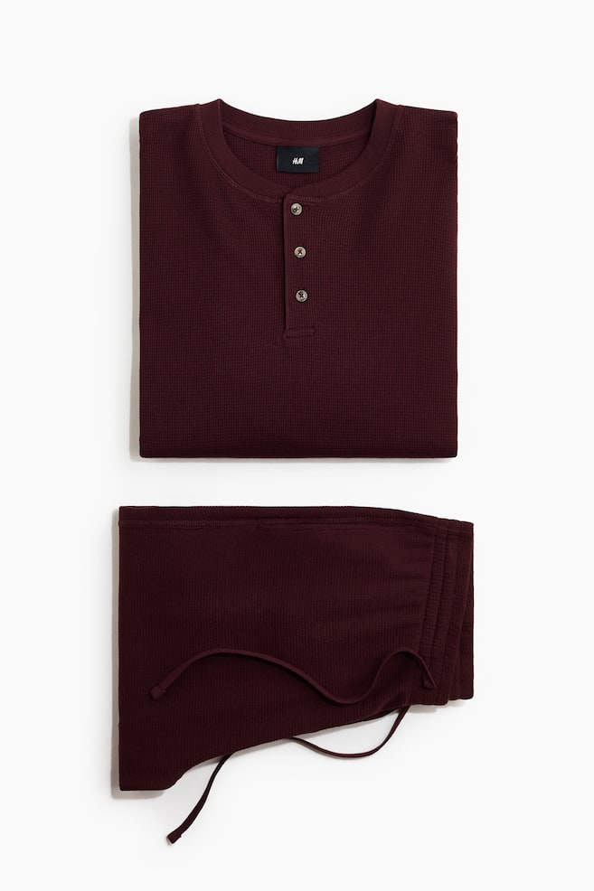 Ein bordeauxfarbenes Loungewear-Set aus gewaffeltem Baumwolljersey liegt gefaltet in einer Flat-Lay-Ansicht und präsentiert ein Henley-Shirt mit geripptem Rundhalsausschnitt und einer Drei-Knopf-Leiste, die über einer passenden Hose mit elastischem Bund und Kordelzug angeordnet ist. - 1