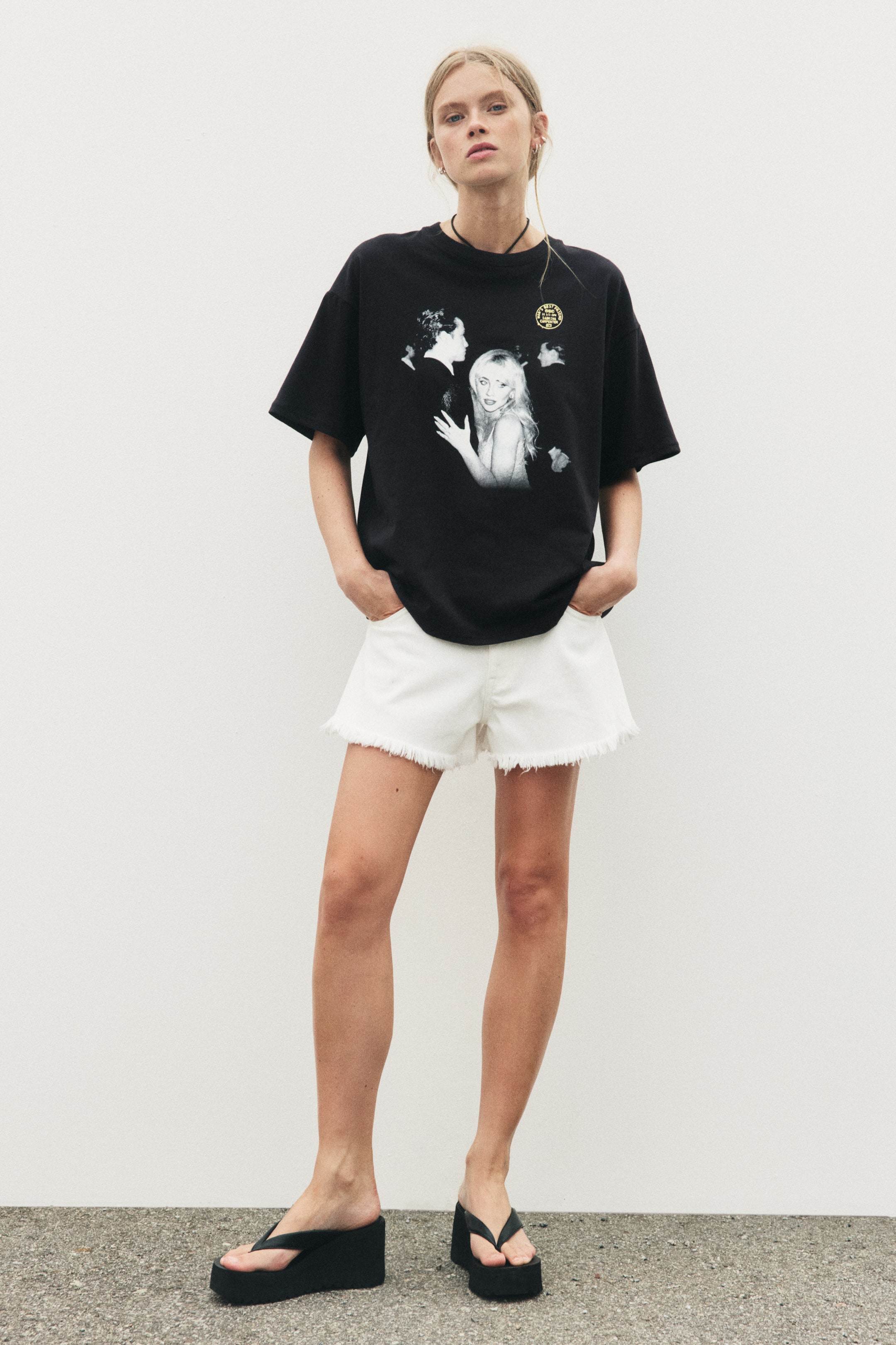Visualizza l'immagine ingrandita: Una t-shirt oversize in jersey di cotone nero è indossata dalla modella. Presenta sul davanti una stampa grafica in bianco e nero raffigurante una donna bionda e altre figure, con un piccolo dettaglio circolare giallo, oltre a un bordo a costine lungo lo scollo e spalle scese. Le mani della modella sono nelle tasche dei pantaloncini, facendo cadere la t-shirt in modo morbido.