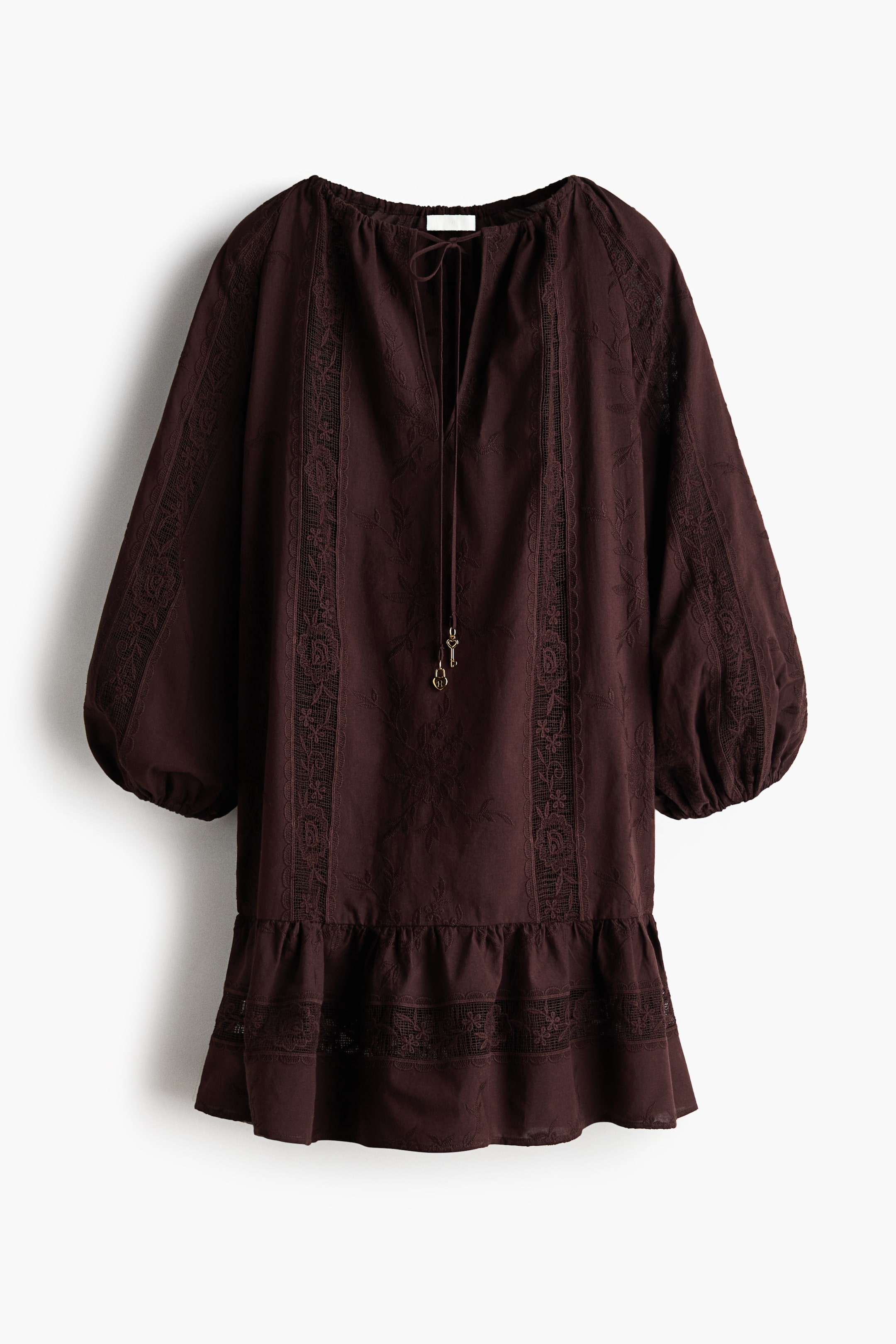Ampliar la imagen: Un vestido túnica corto, de color marrón oscuro y de corte holgado, presenta su parte frontal, confeccionado en un tejido de algodón con motivos florales bordados e inserciones verticales de encaje. Cuenta con un escote redondo con cordón ajustable y una abertura en forma de V con pequeños dijes de llaves, mangas largas tipo raglán con puños elásticos, y una costura fruncida en las caderas que crea un dobladillo ligeramente acampanado.