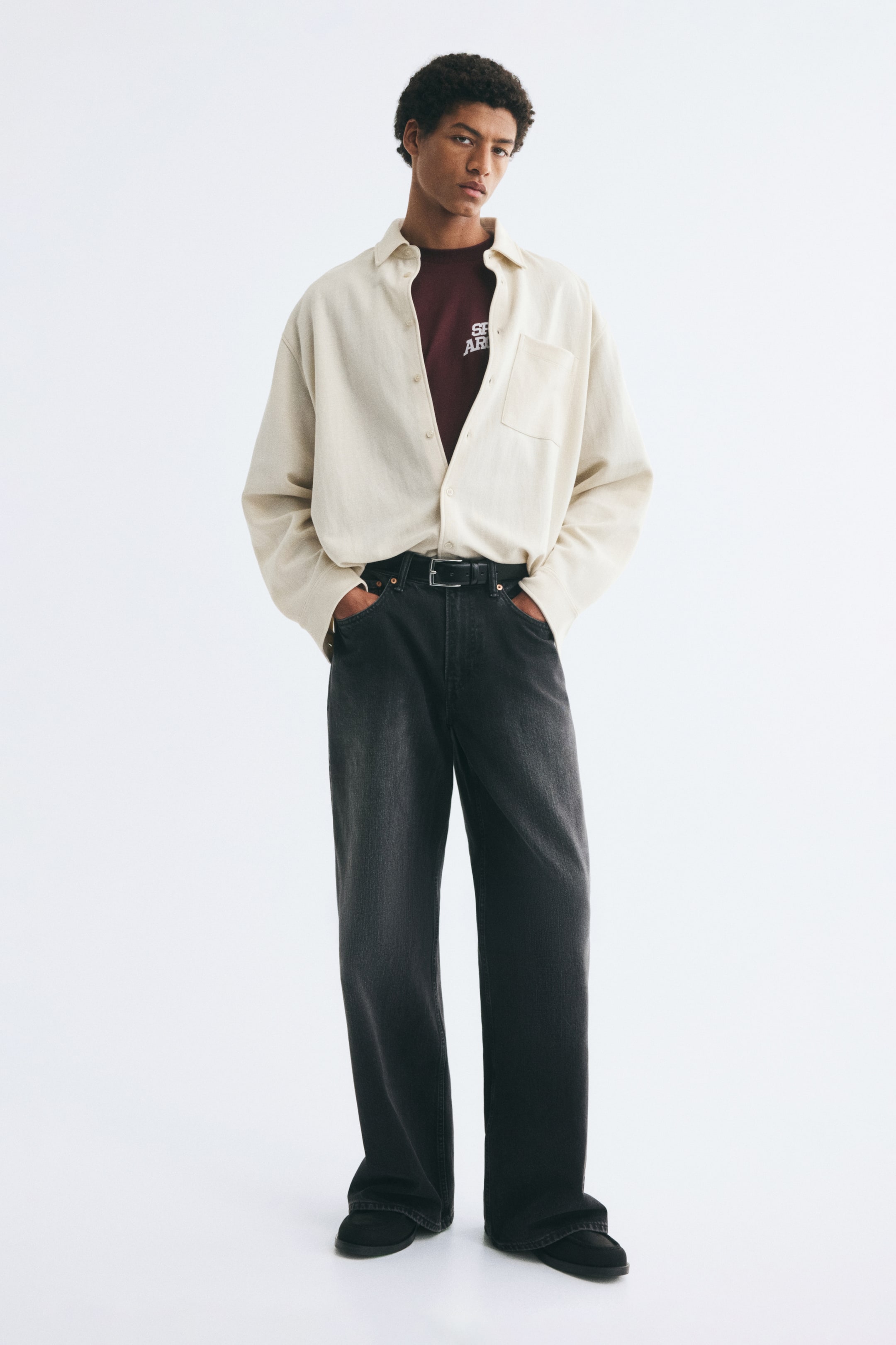 View larger image: A dark burgundy T-shirt featuring a white printed graphic on the chest is worn by the model. The product is styled tucked into dark grey wide-leg jeans and layered under an unbuttoned cream-coloured long-sleeve shirt.