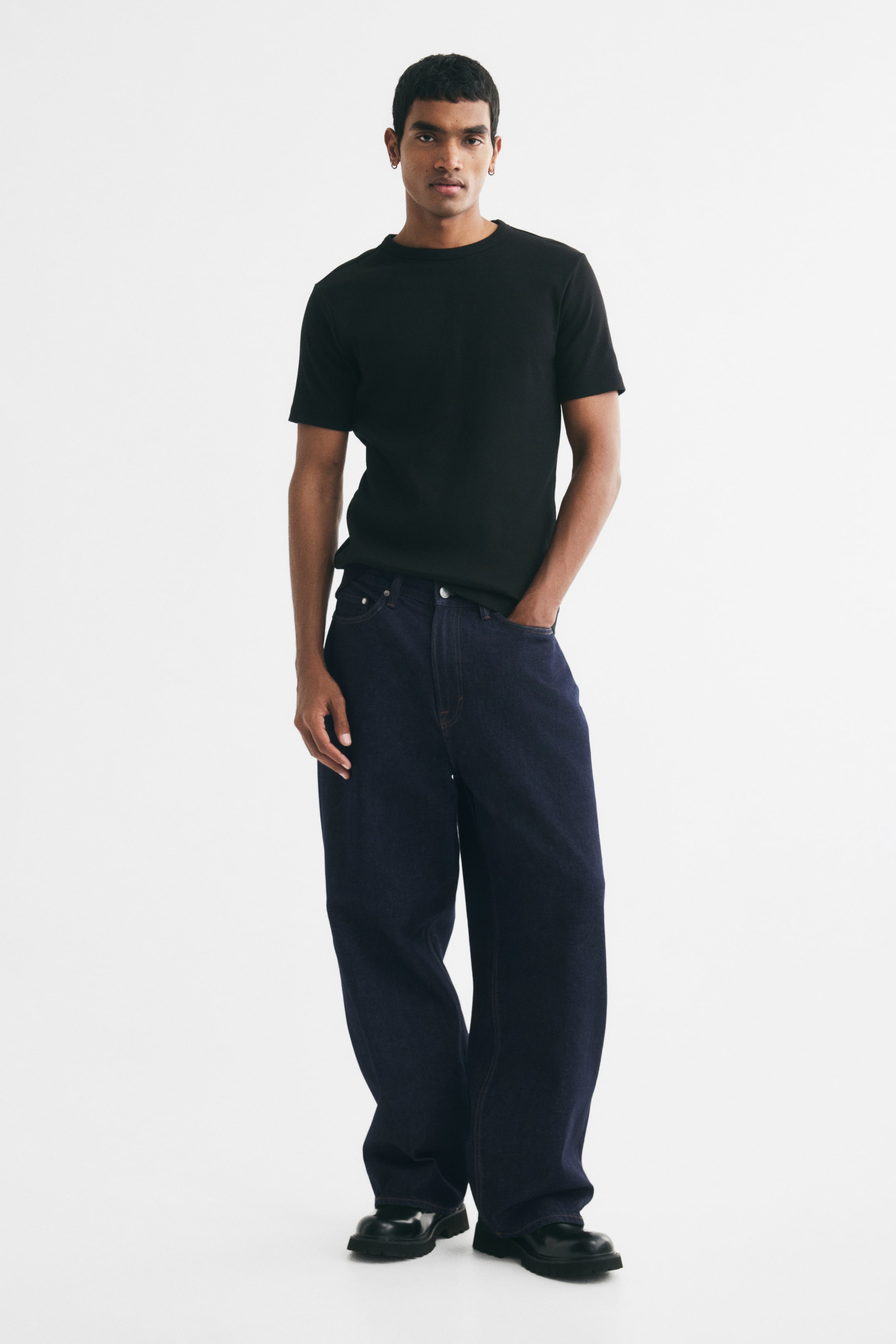 View larger image: The model is wearing dark blue denim jeans with a baggy, rounded-leg fit, standing with one hand in a pocket. The jeans feature a regular waist and stack at the ankles over black shoes, styled with a black short-sleeve T-shirt.