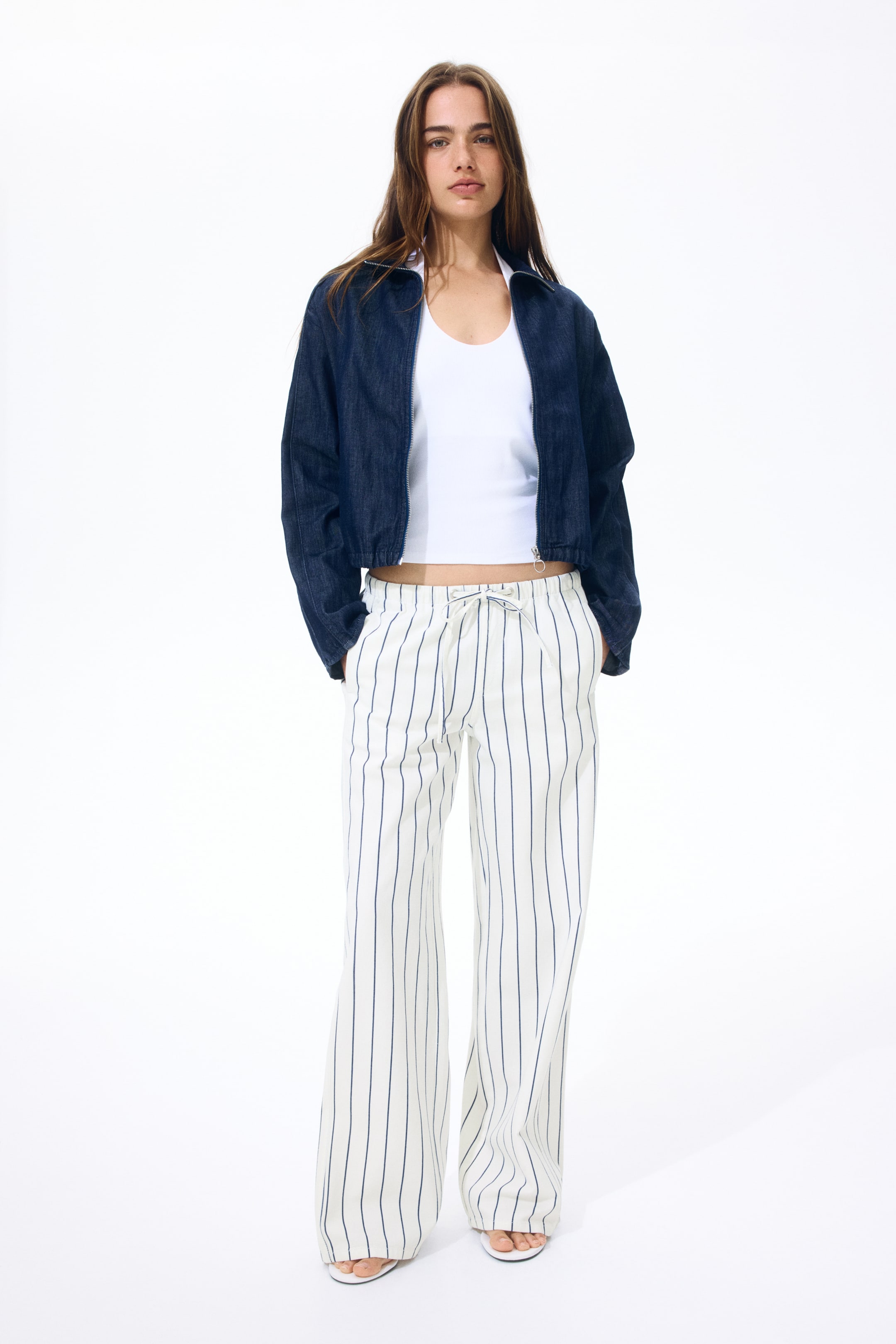 Größeres Bild ansehen: Eine dunkelblaue Denim-Cropped-Jacke mit einem Kragen und einem Frontreißverschluss wird über einem weißen Tanktop mit Rundhalsausschnitt getragen. Weiße Hosen mit weitem Bein präsentieren vertikale marineblaue Streifen und einen Kordelzug in der Taille, während weiße offene flache Sandalen das nach vorne gerichtete Ensemble vervollständigen.