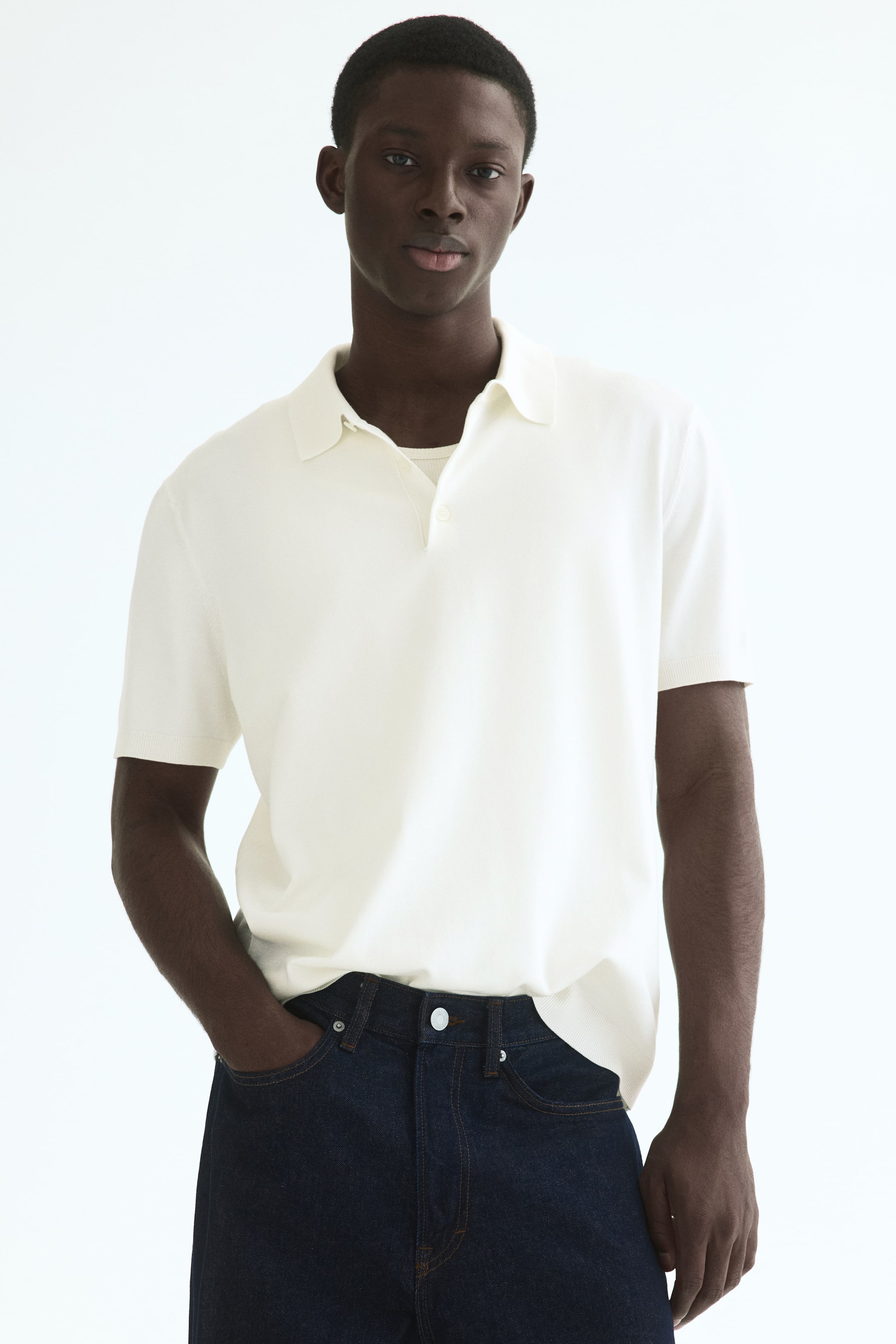 View larger image: A cream fine-knit short-sleeved polo shirt is showcased facing forward, featuring a classic collar and a two-button placket. It is subtly layered over a light undershirt, with its hem resting over dark jeans.