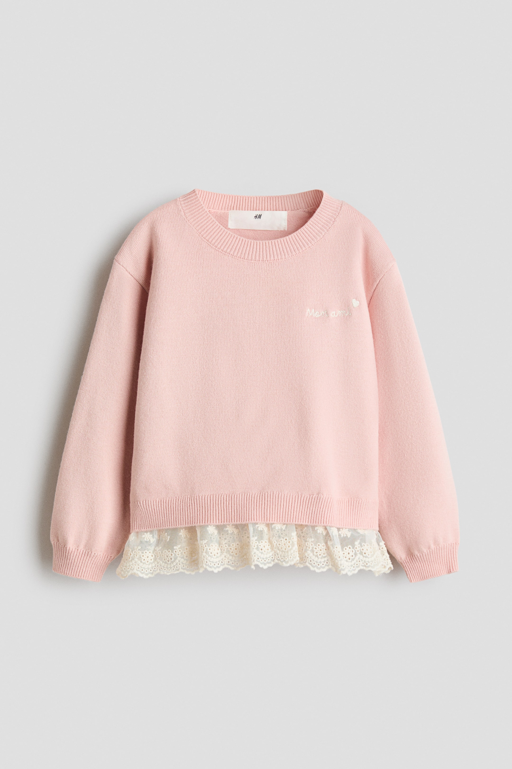View larger image: A pink fine-knit sweater is shown from the front, featuring a round ribbed neckline, dropped shoulders, and a small white embroidered text detail on the chest. A sewn-in, sheer cream broderie anglaise layer with scalloped edges extends from beneath the ribbed hem.
